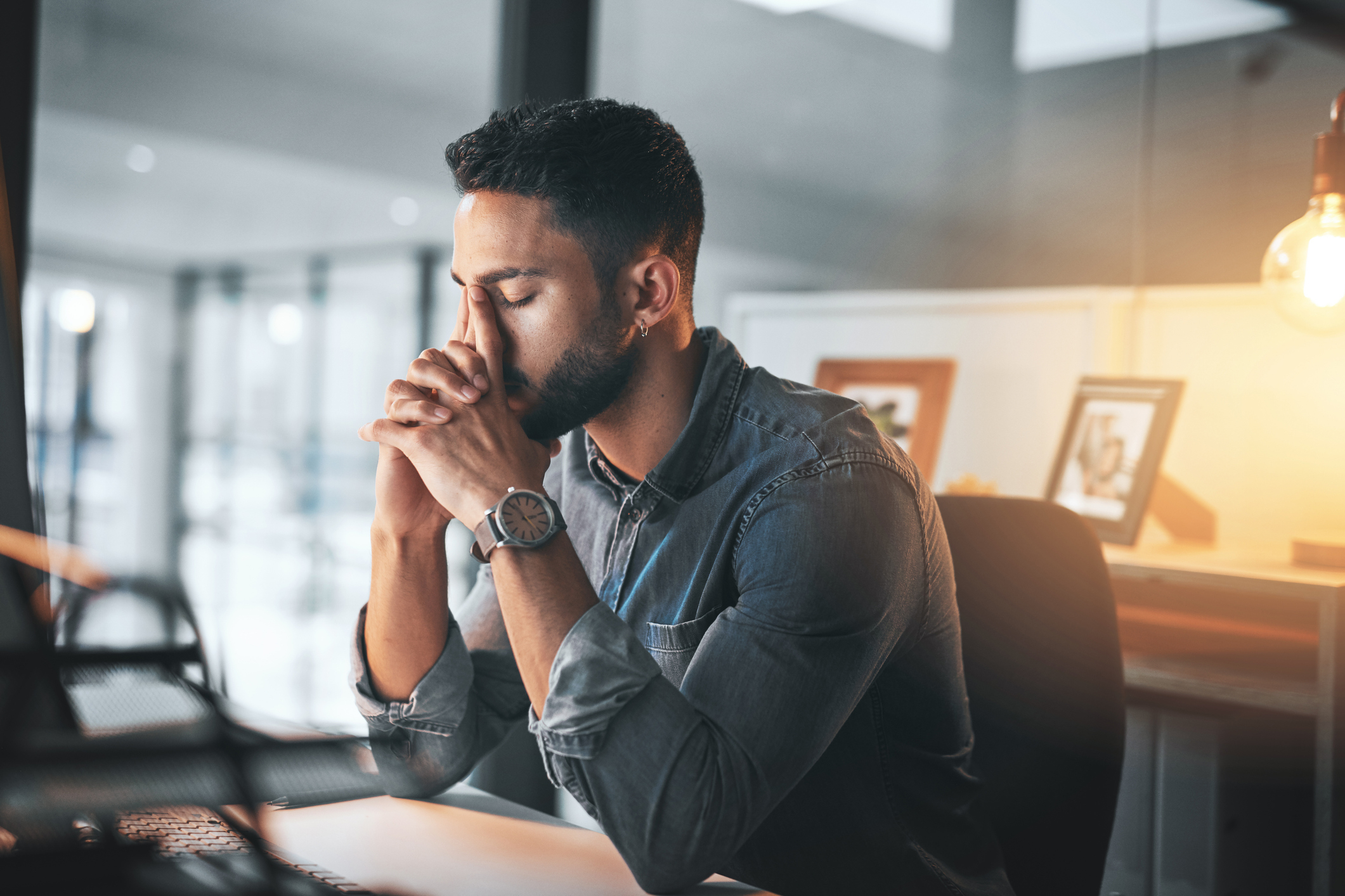 A man sits at a desk, eyes closed and face resting on clasped hands, appearing stressed or deep in thought