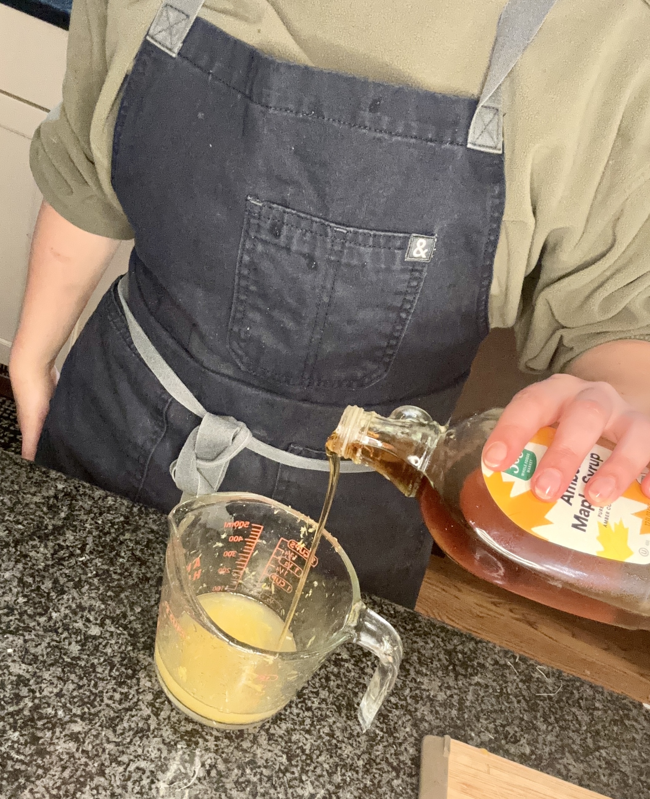 Person pouring dressing over a large bowl containing vegetables, roasted potatoes, and herbs in a kitchen. Only the hands and torso are visible