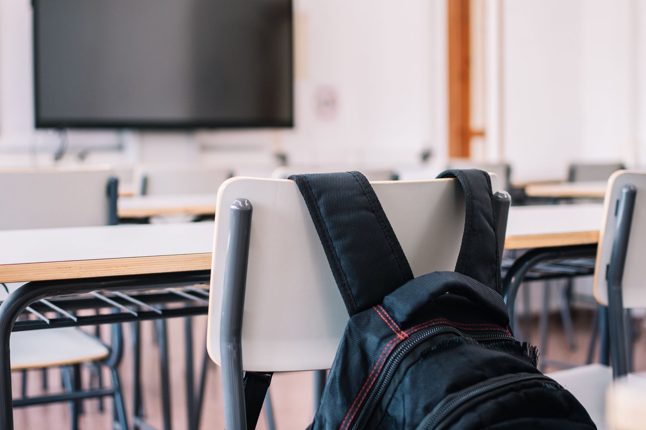 A close-up of a classroom with empty chairs and desks, featuring a backpack hanging over the back of a chair