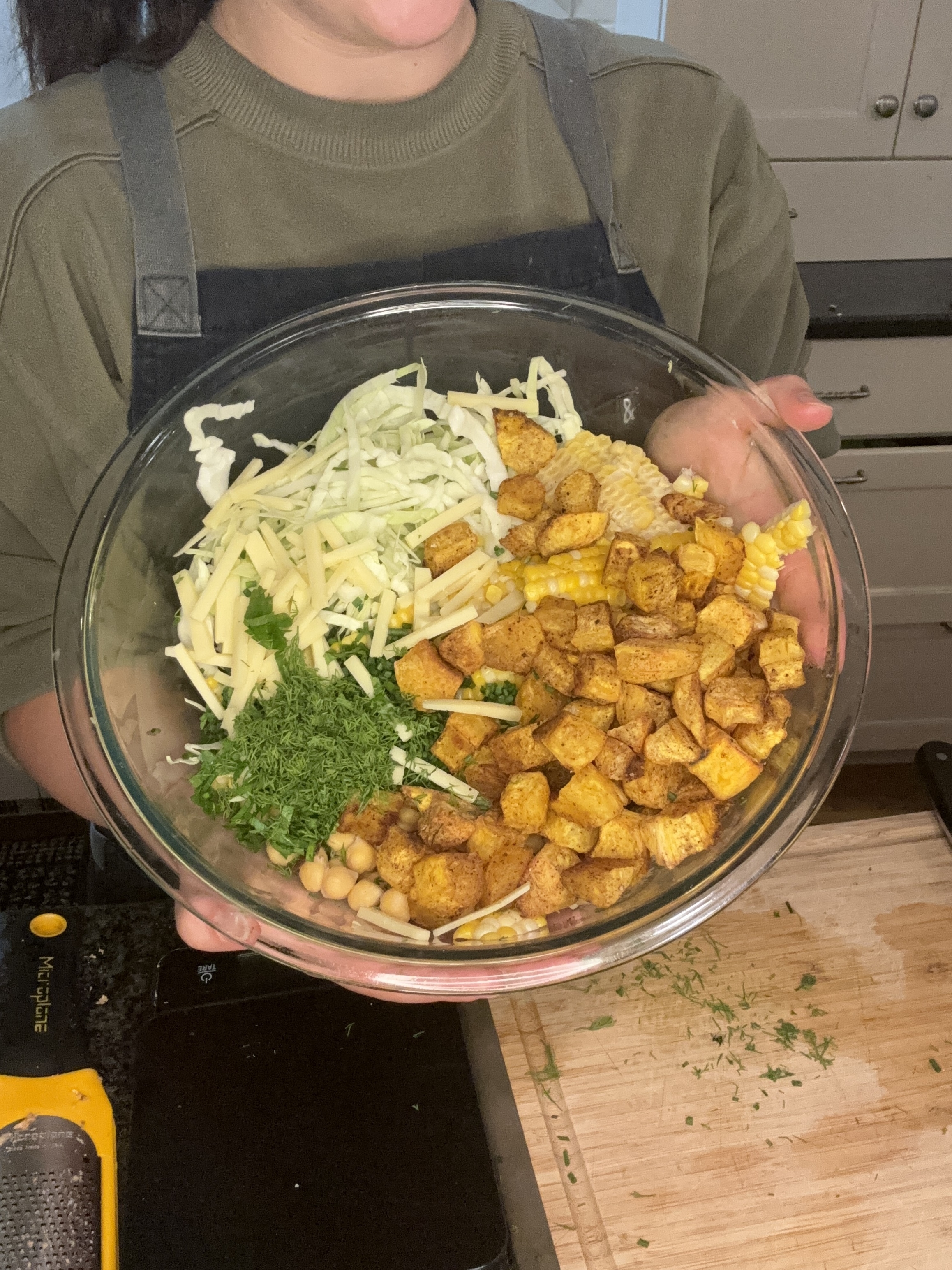 Person holding a bowl with chopped cabbage, grated cheese, roasted potatoes, corn, parsley, and chickpeas in a kitchen