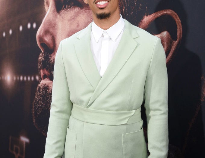 Man in a stylish suit on a red carpet in front of a movie poster depicting a close-up of another man's face. Name is not provided