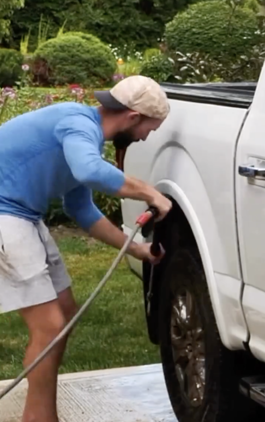 A man in casual attire washes the tire of a white pickup truck in a garden with lush greenery in the background
