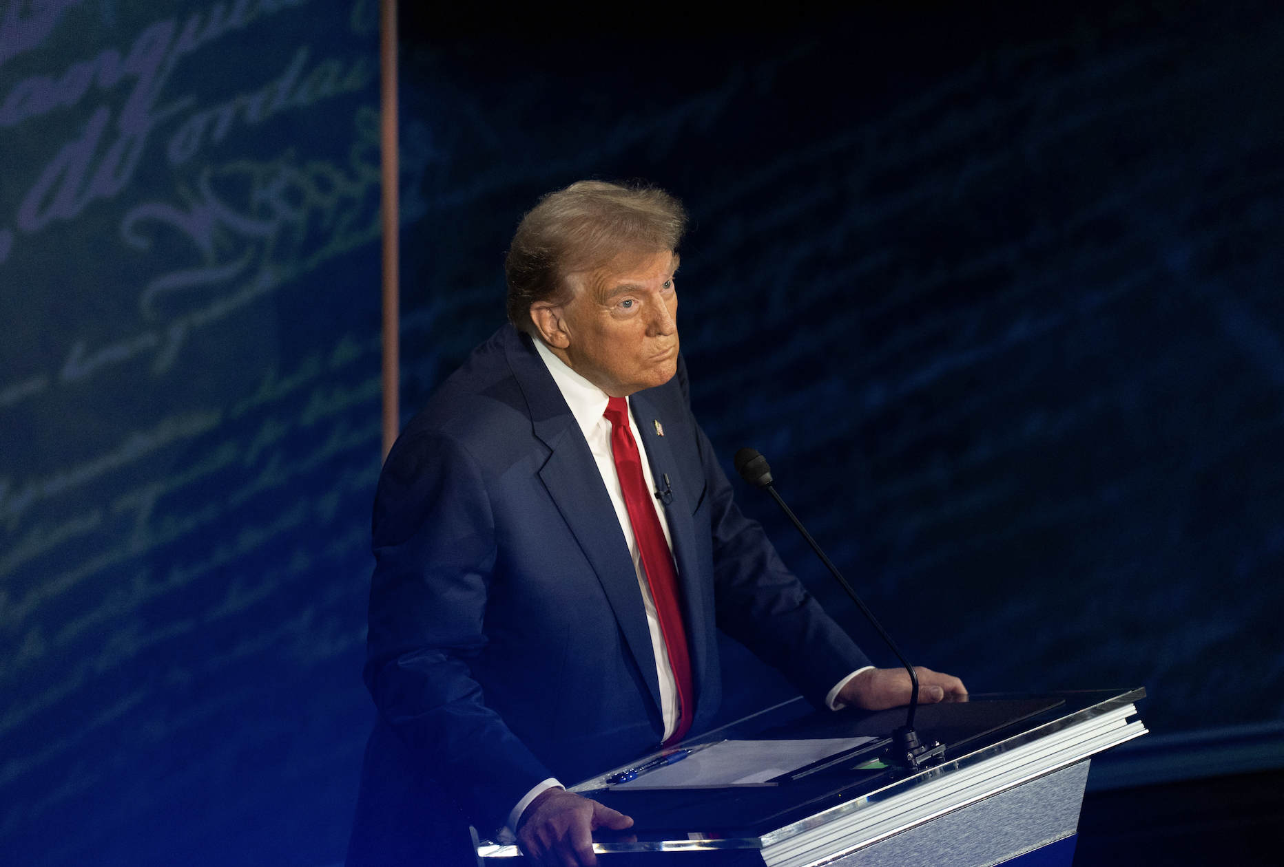 Donald Trump stands at a podium during a debate, wearing a suit with a red tie. Handwritten words are visible in the background