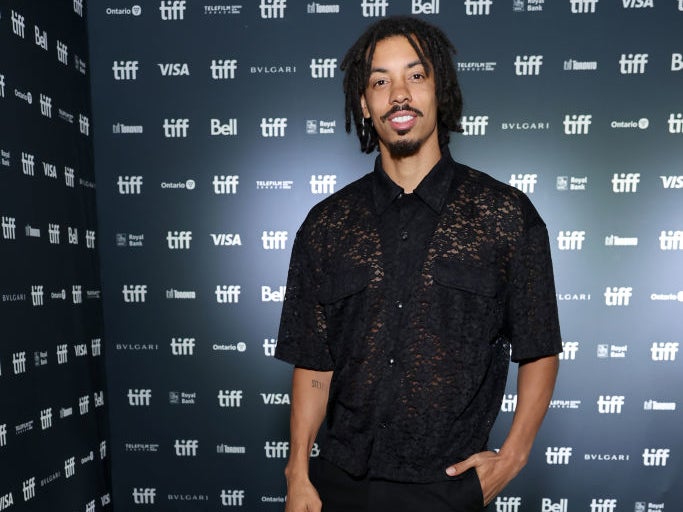 Melvin Gregg posing on the red carpet at TIFF, wearing a sheer black shirt and black pants, with a step-and-repeat background featuring festival sponsors
