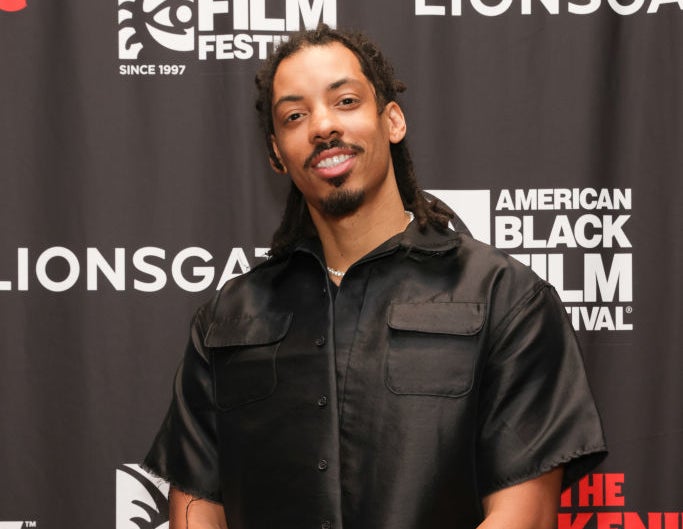Man posing in front of an American Black Film Festival banner wearing a black short-sleeve shirt and blue jeans