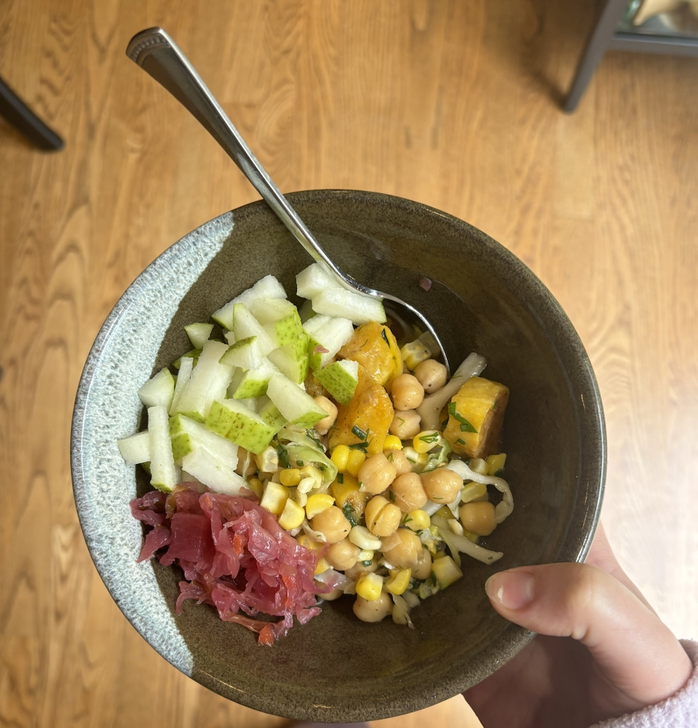 A person holds a bowl of chickpeas, diced pears, squash, pickled red onion, and corn with a spoon on a wooden surface