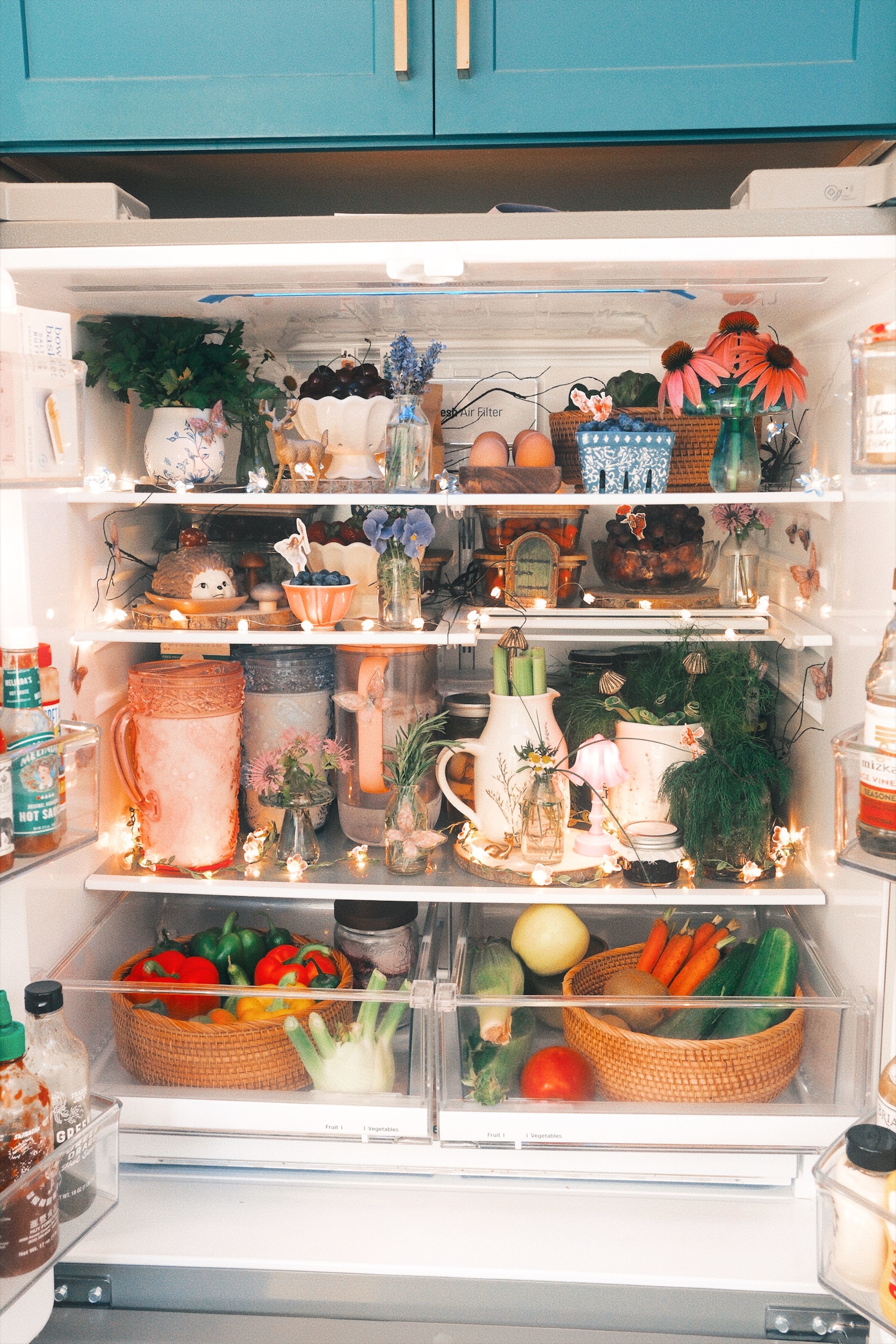 A neatly organized fridge decorated with various plants, flowers, and fairy lights; fresh produce, eggs, and jars are also arranged on the shelves