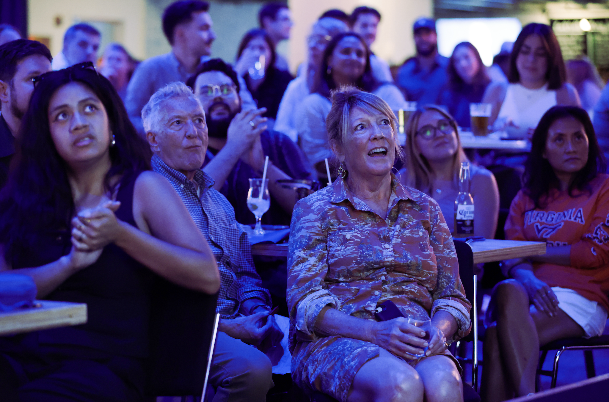 A group of people, including a woman in a patterned dress, watch an event with expressions of anticipation and excitement
