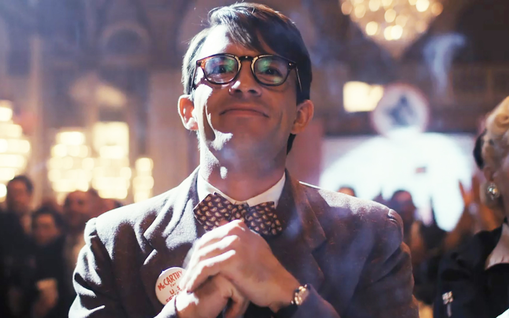 A person with glasses and a bow tie smiles and clasps their hands at a formal event. They are wearing a suit with a name badge. The background is a blurred crowd