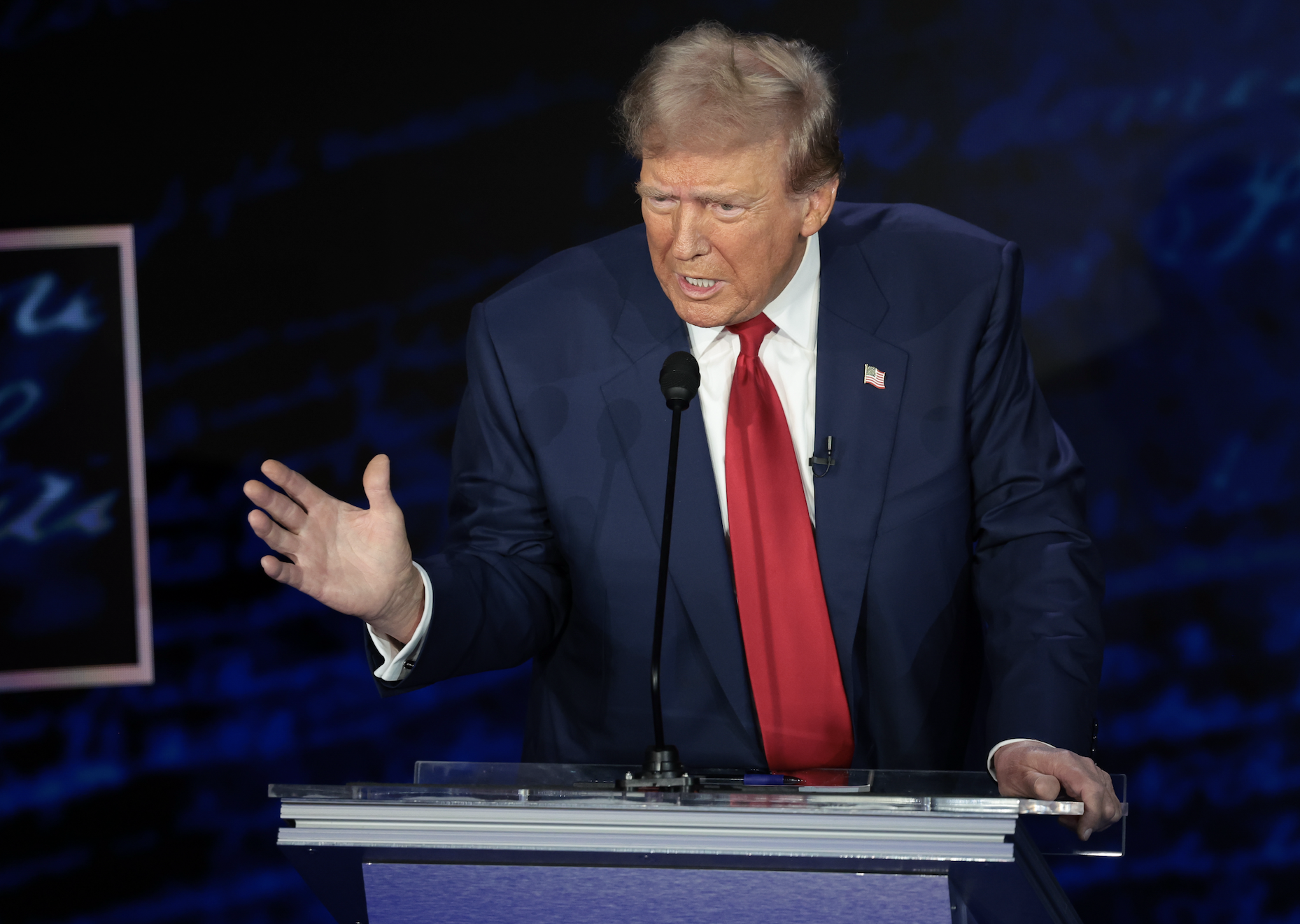 Donald Trump stands at a podium, gesturing with his hand while speaking at a debate or public event