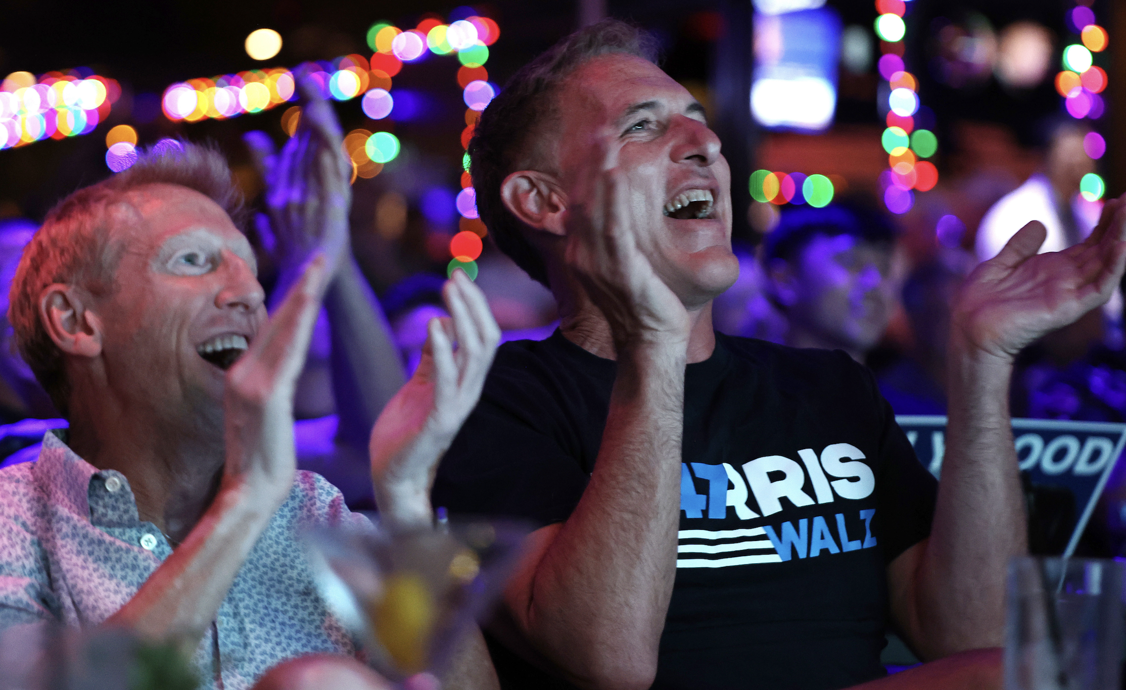 Two enthusiastic men in a lively setting clap and cheer, with colorful lights in the background. The man on the right wears a shirt with the text &quot;Harris Waltz.&quot;