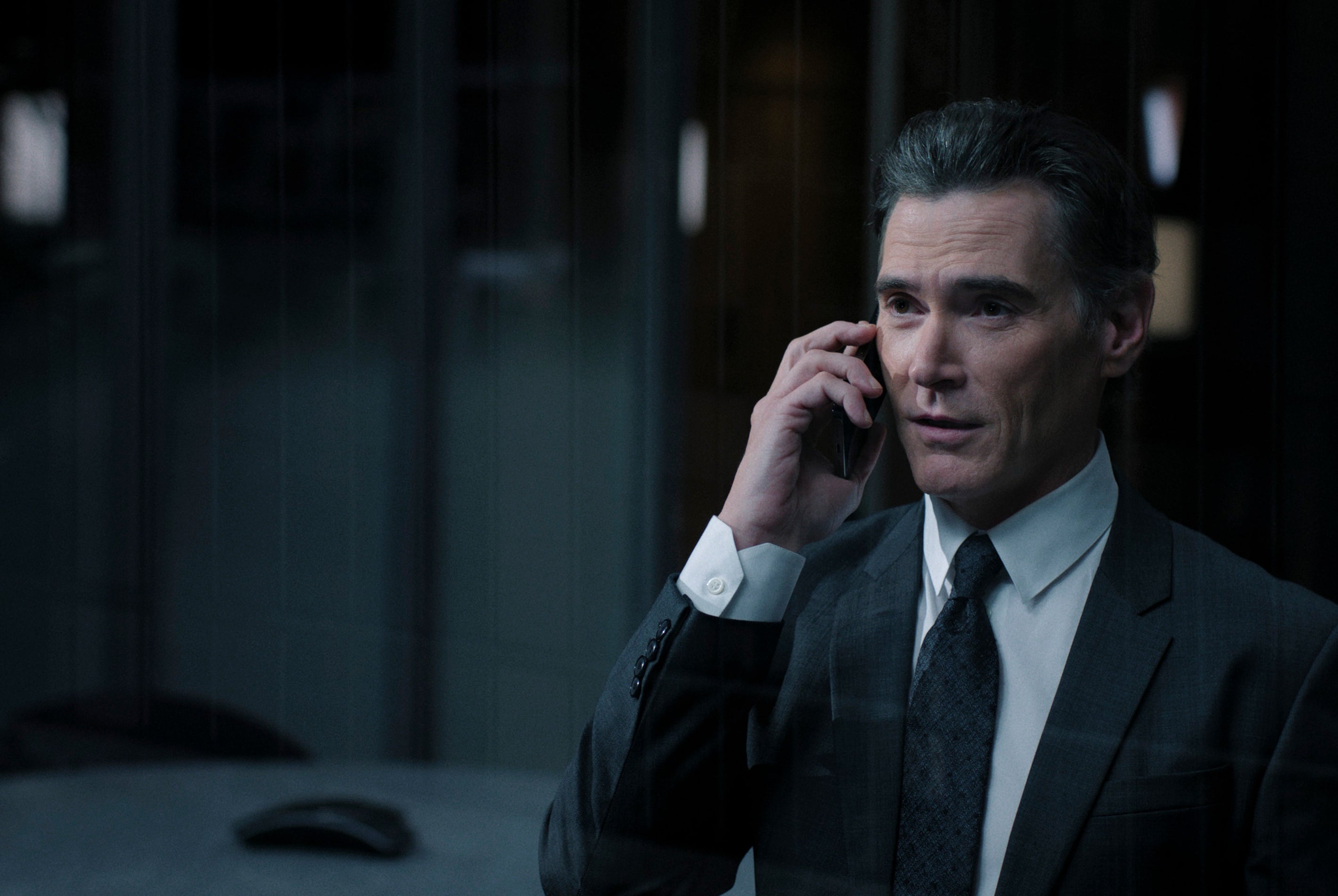 Billy Crudup in a suit, on a phone call, in a dimly lit office setting