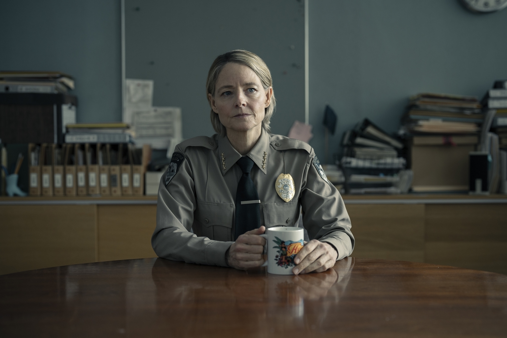 Jodie Foster, dressed in a sheriff's uniform, sits at a wooden table holding a mug, with an office background filled with files and binders