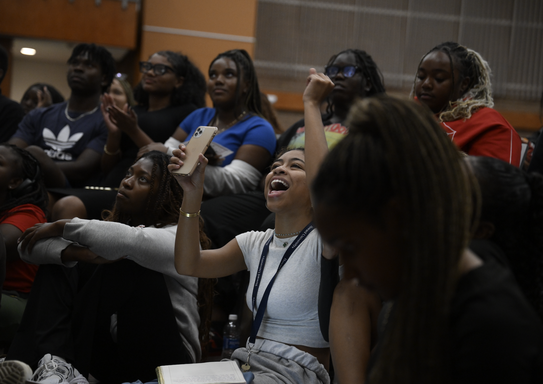 Students in a lively classroom setting, one student, holding a phone, raises her fist excitedly while others around her appear engrossed and attentive