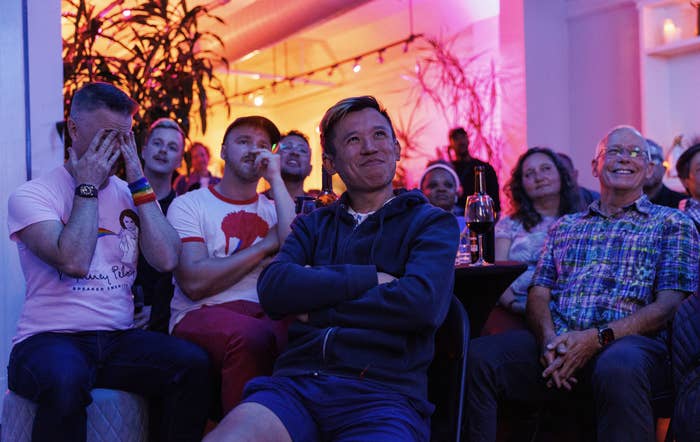 Group of people, including one man with head in hands and another smiling with arms crossed, seated and watching something off-camera in a cozy setting with indoor plants