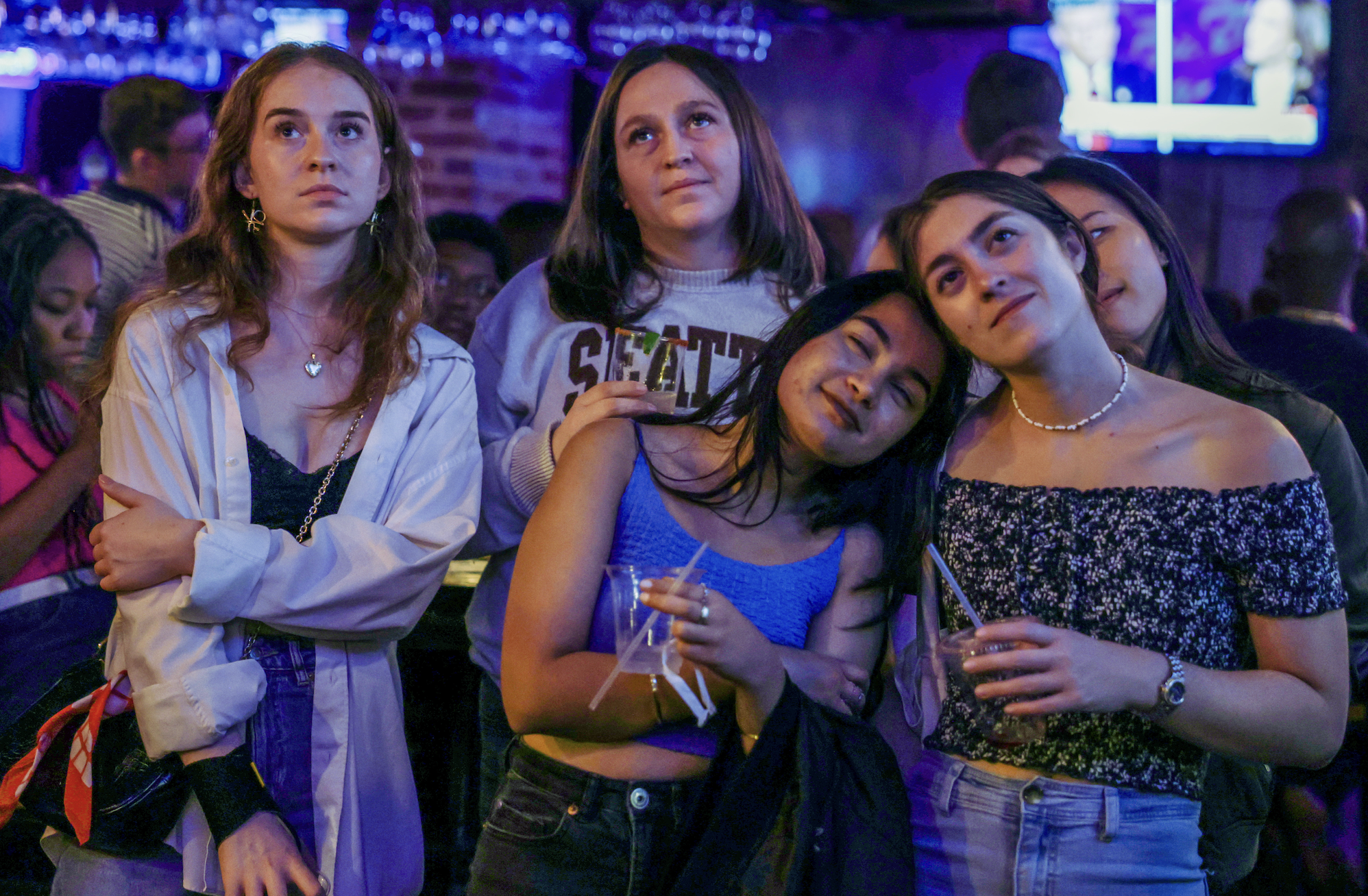 Five women in casual attire pose together in a dimly lit bar, with two TV screens in the background showing blurred content
