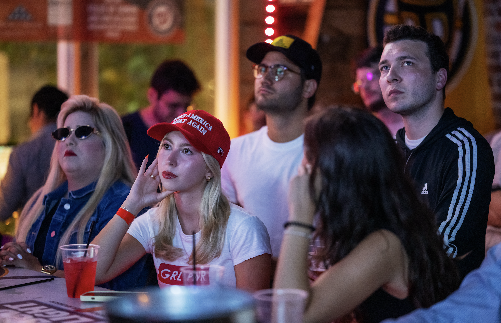 Group of people at a bar watching an event on TV. One woman wears a red &quot;Make America Great Again&quot; hat with her hand on her face. Names unknown