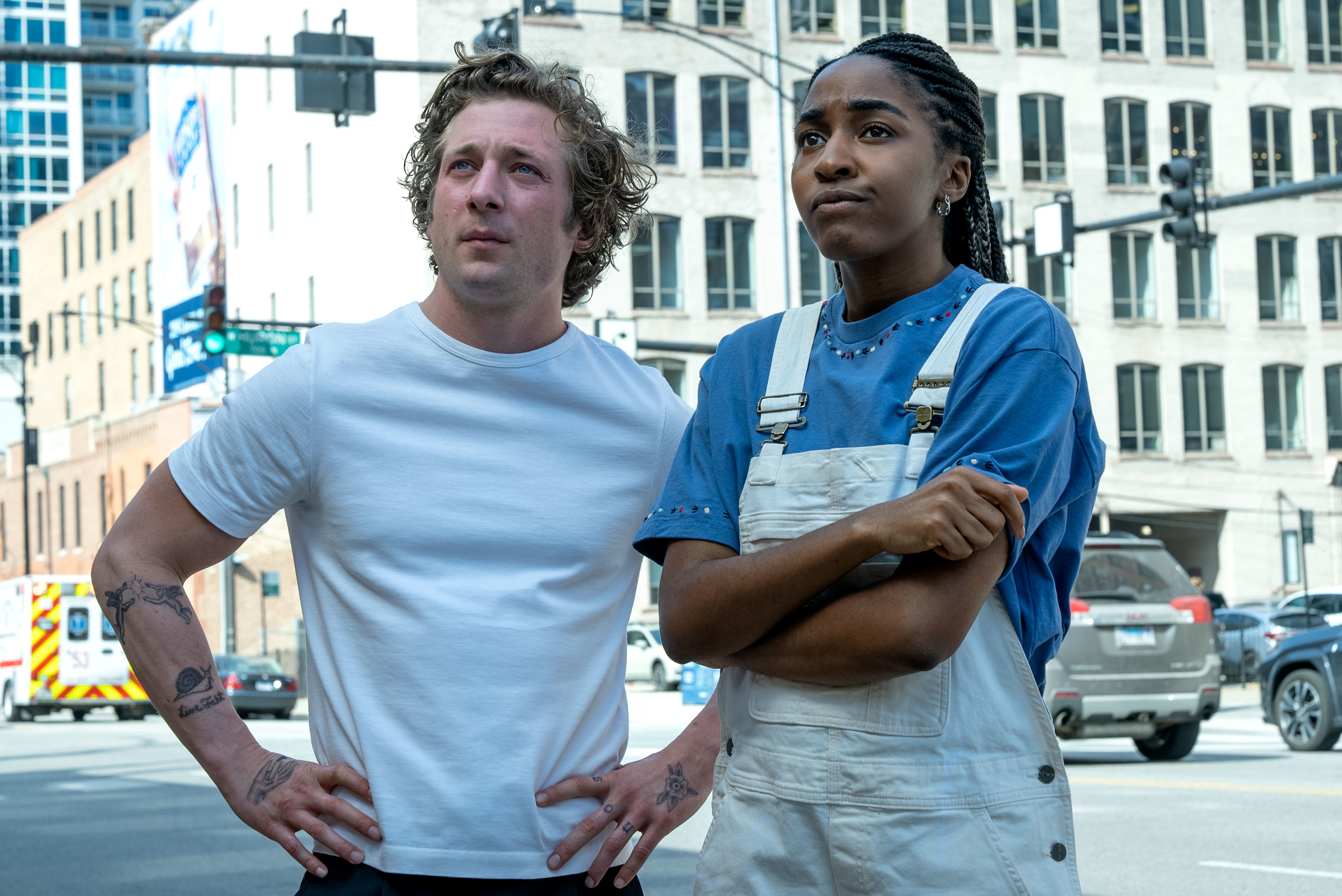 Jeremy Allen White in a t-shirt and Ayo Edebiri in a t-shirt and overalls on a city street, looking into the distance in a scene from &quot;The Bear&quot;