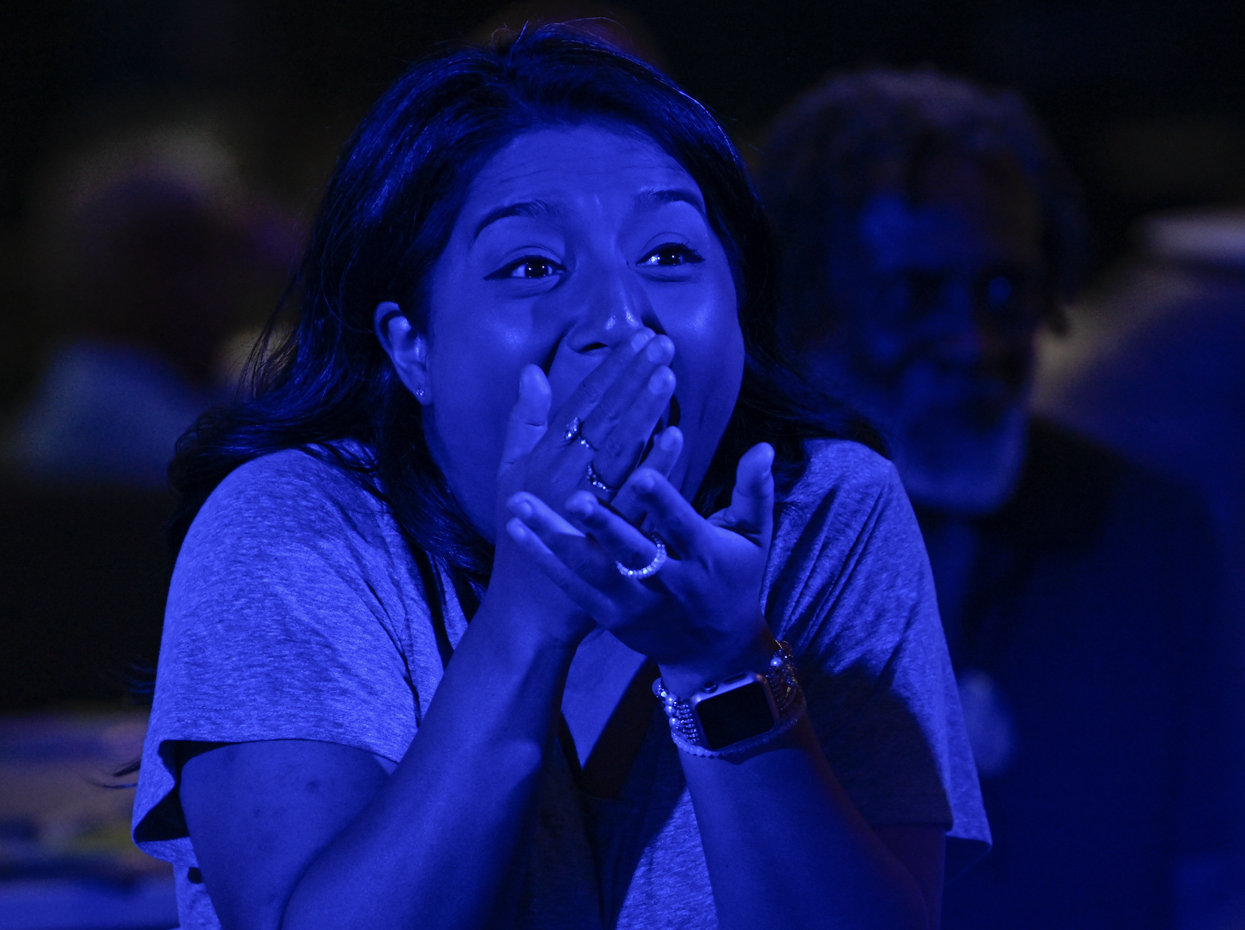 Woman with surprised expression covers mouth with both hands. Background blurred and has an elderly man
