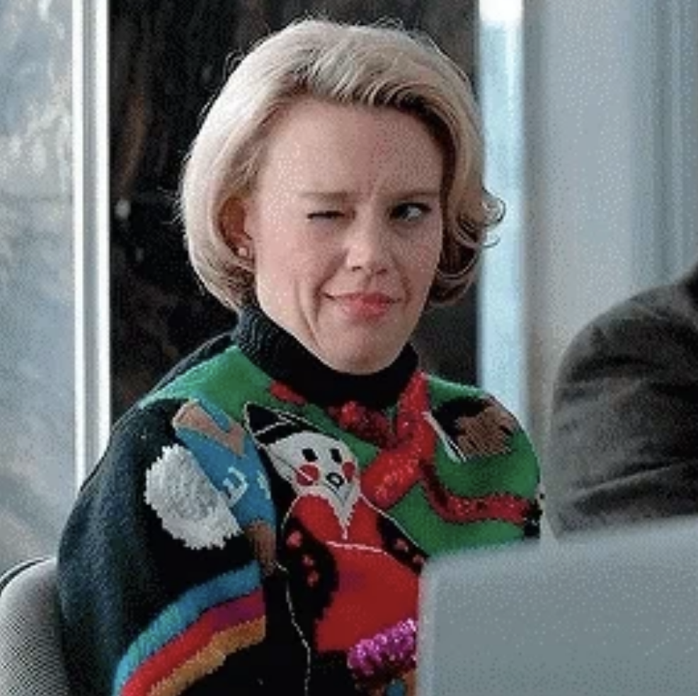 Kate McKinnon winks while wearing a festive Christmas sweater featuring a snowman and a large bow