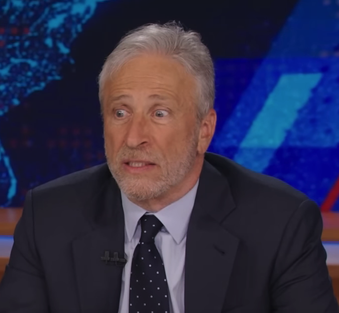 Jon Stewart, wearing a suit and polka dot tie, looking surprised during a TV broadcast