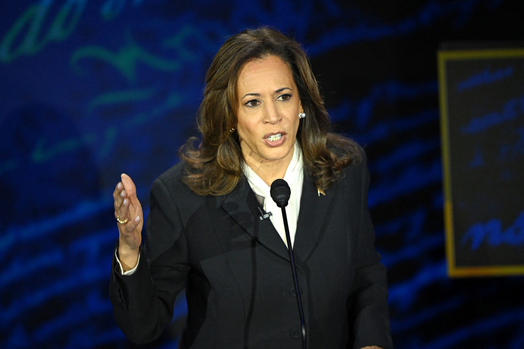 Kamala Harris speaking at a podium during an event. She is wearing a dark formal suit and gesturing with her right hand