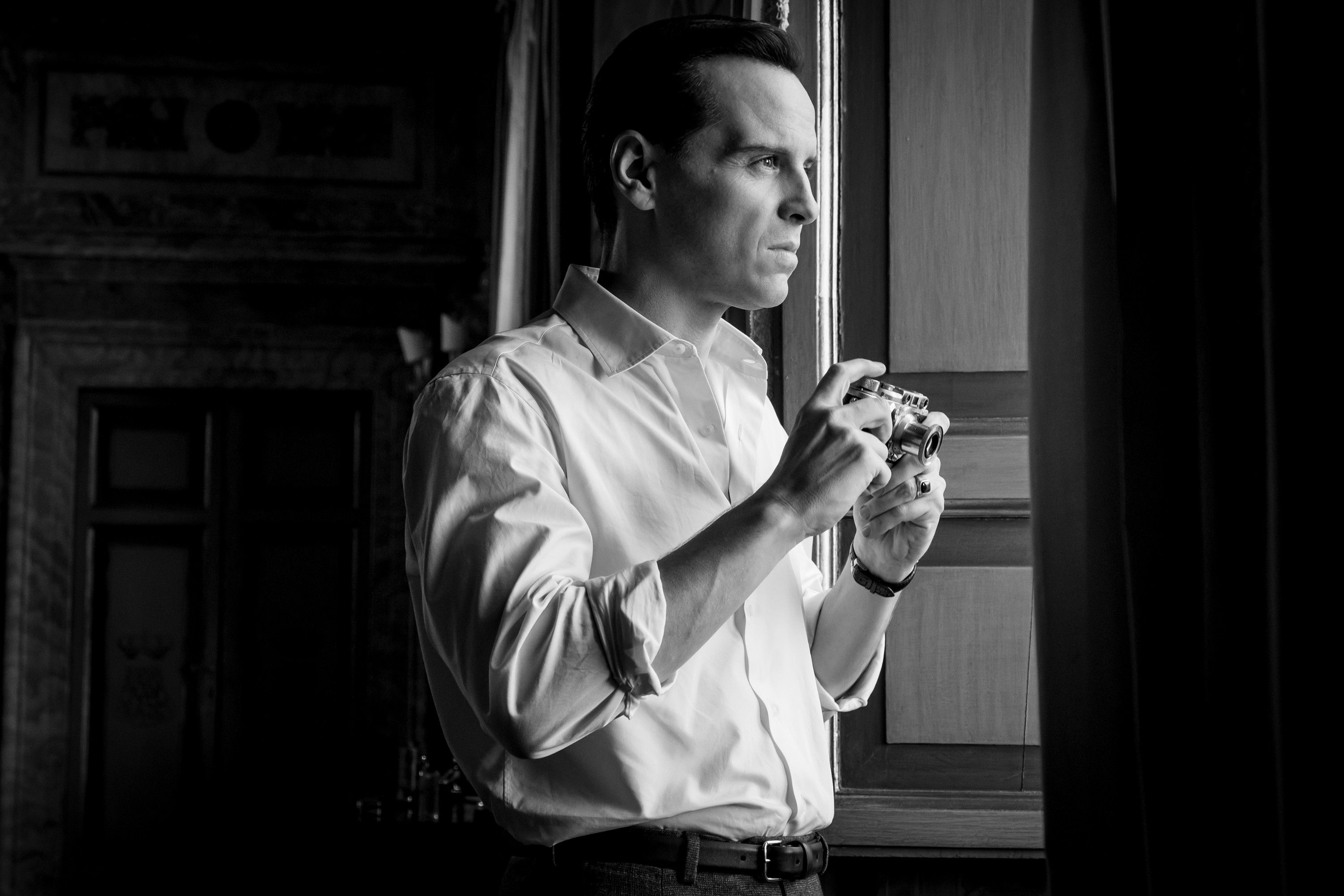 Andrew Scott stands by a window holding a camera, wearing a button-up shirt in a scene from a TV show or movie