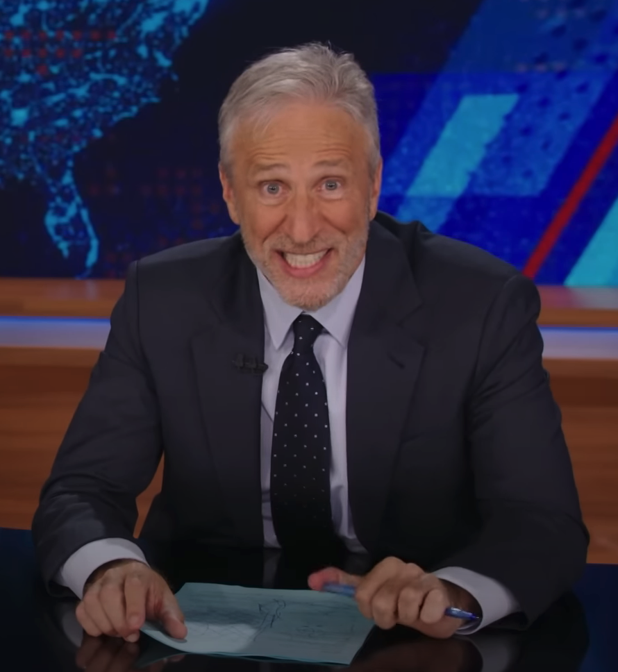 Jon Stewart, seated at a news desk, holding paper and smiling while wearing a suit and polka dot tie