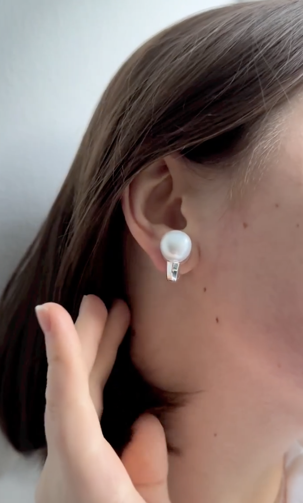A close-up of a person with short brown hair wearing a single pearl earring. They are touching their ear lightly