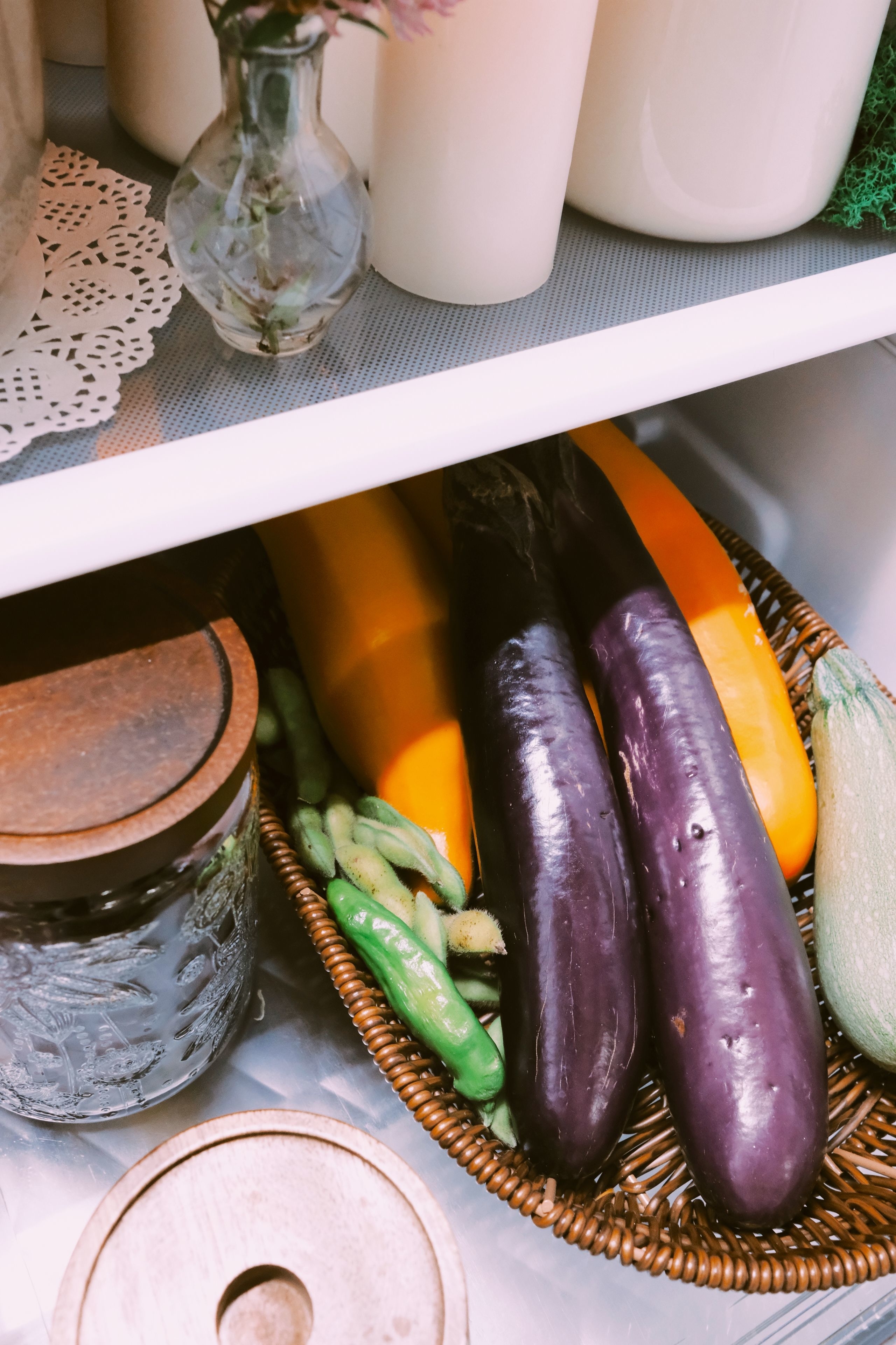A refrigerator shelf contains a wicker basket of eggplants, zucchini, and string beans. Next to the basket are jars and a small vase with flowers