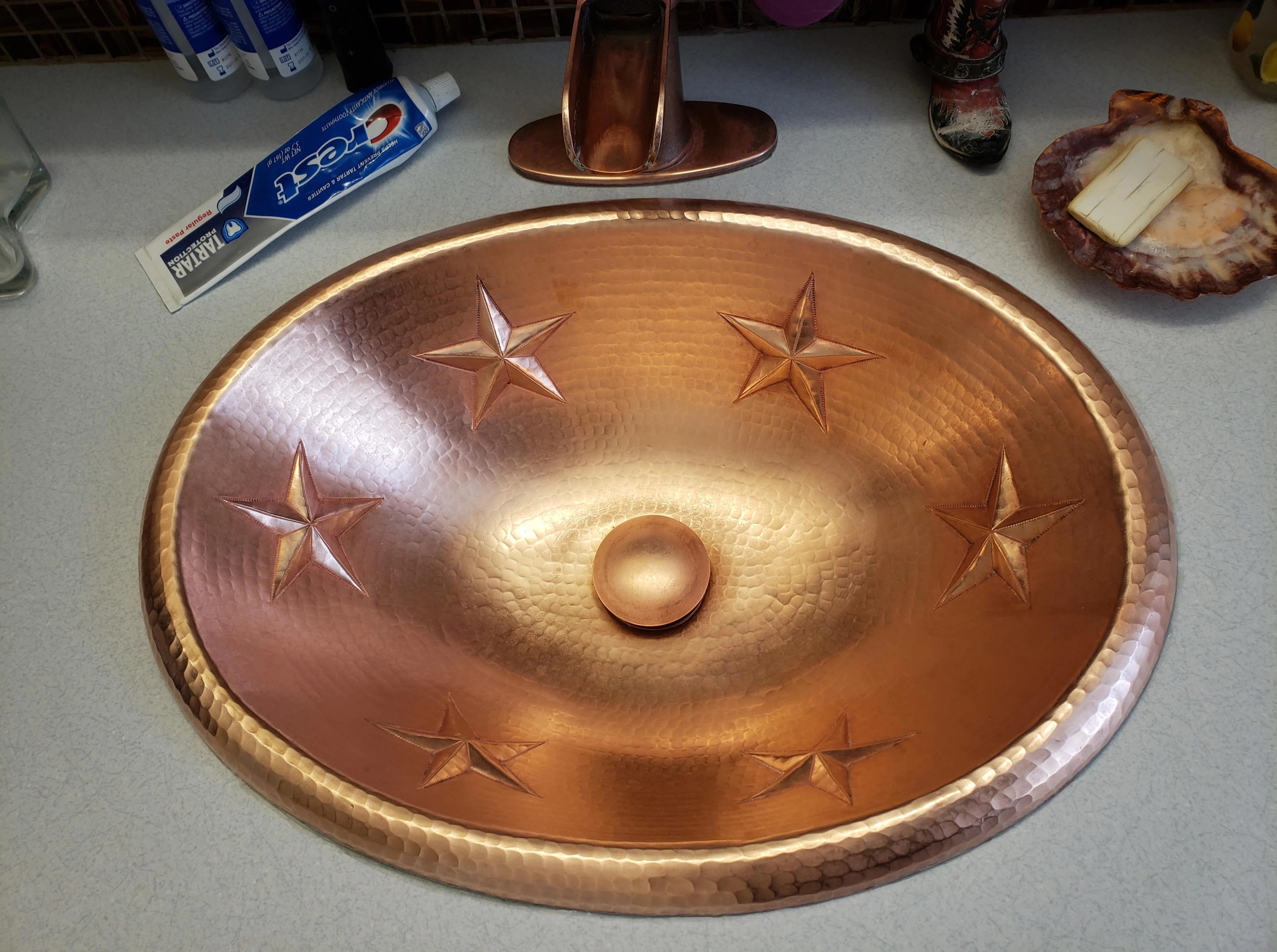 A copper sink with embossed star patterns next to a faucet, a tube of Colgate toothpaste, and a bar of soap in a dish on the counter