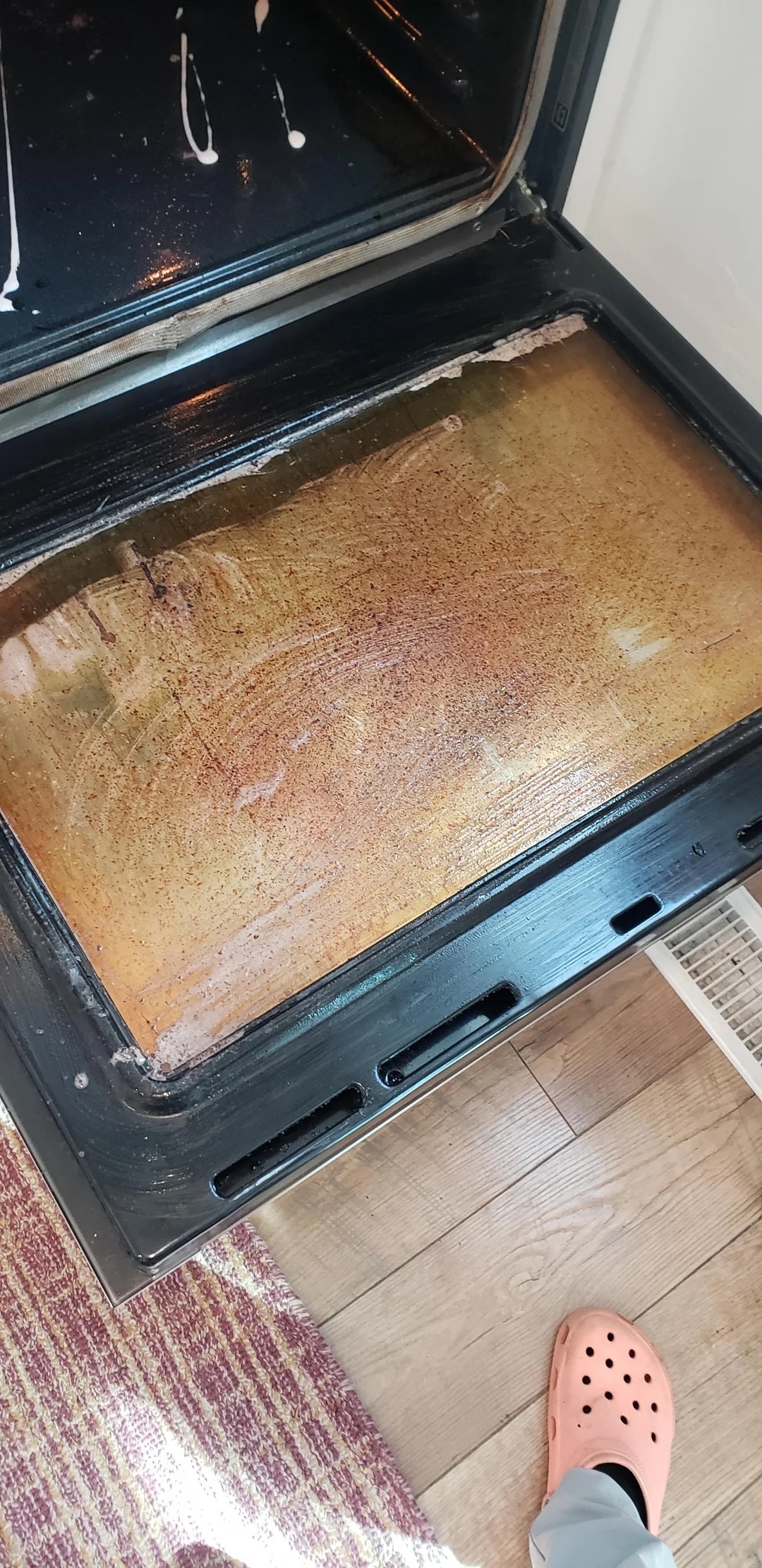 Open oven door with a visible dirty interior. A person's foot wearing a Crocs shoe is visible on a wooden floor next to the oven