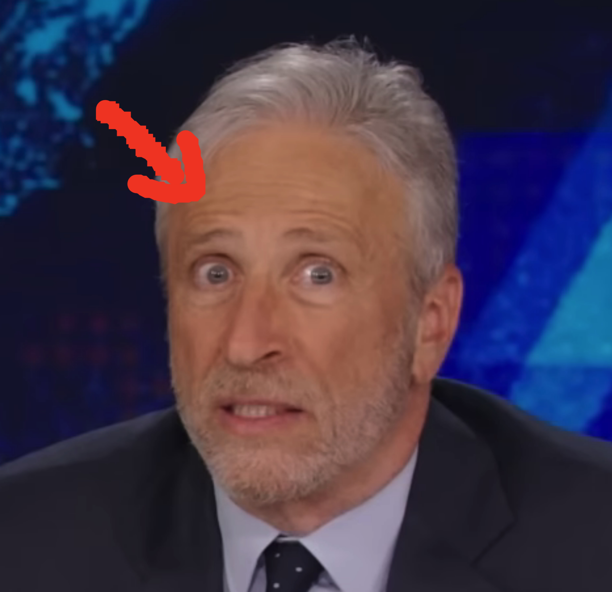 Jon Stewart on a TV set, appearing surprised. He’s wearing a dark suit jacket, white shirt, and polka-dot tie. Background shows a digital map display
