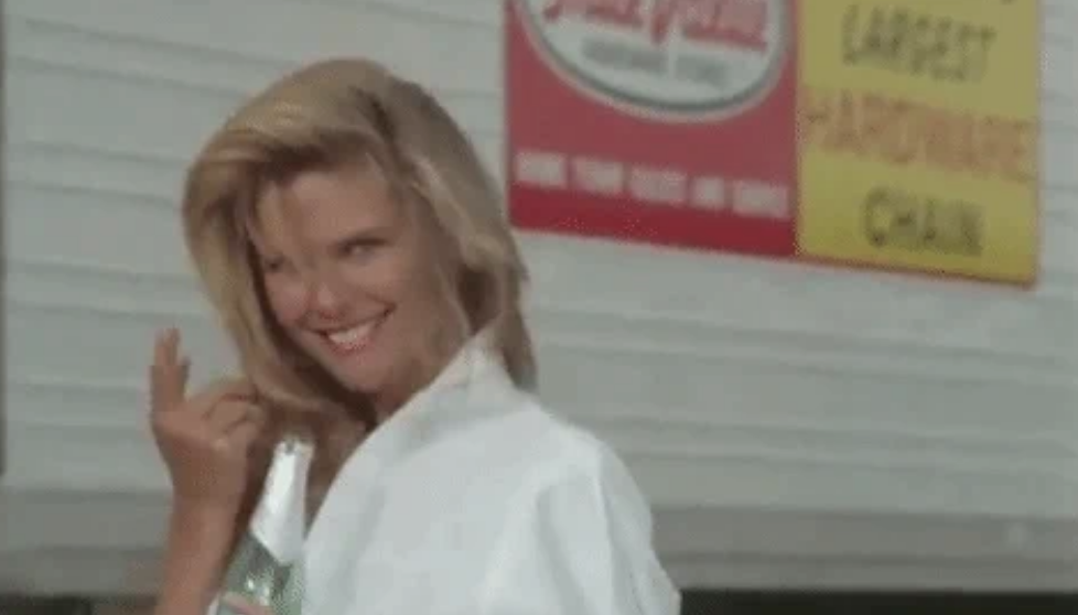 A woman smiling and holding a beverage bottle outside, with a sign reading &quot;Largest Carpenters Chain&quot; in the background