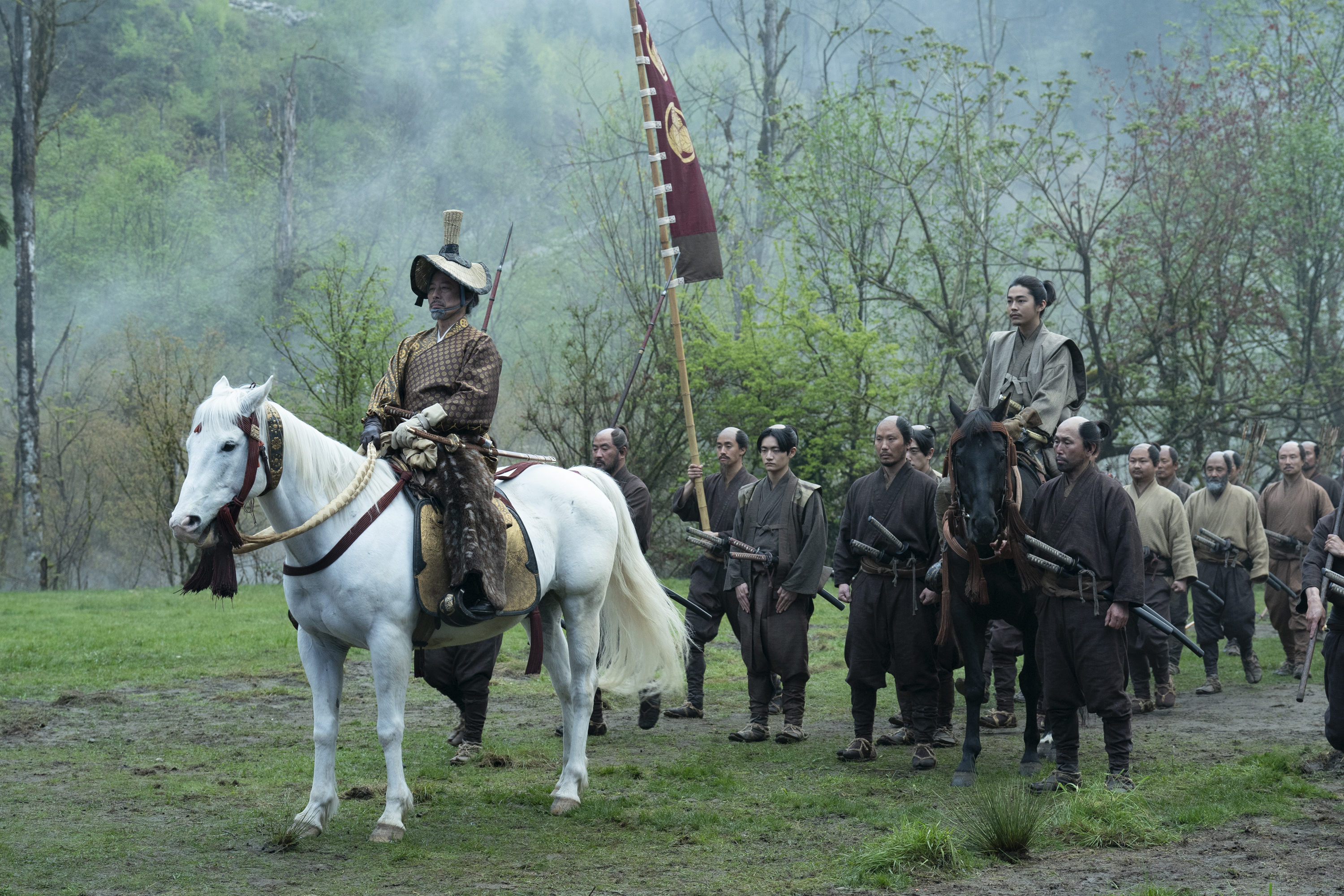 Cast members of &quot;Shogun&quot; dressed in traditional samurai attire, led by a character on a white horse in a forest setting