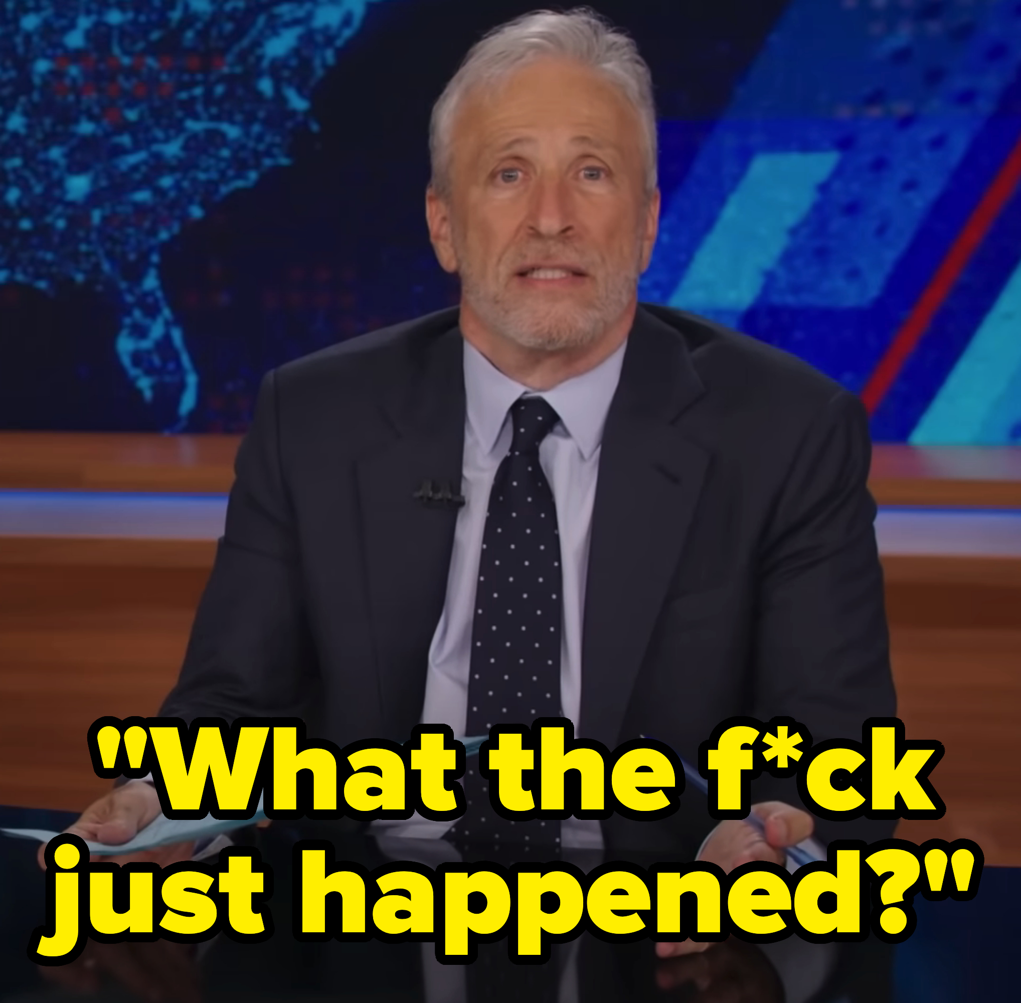 Jon Stewart on a news set, holding papers and a pen, wearing a dark suit and a polka dot tie, speaking to the camera