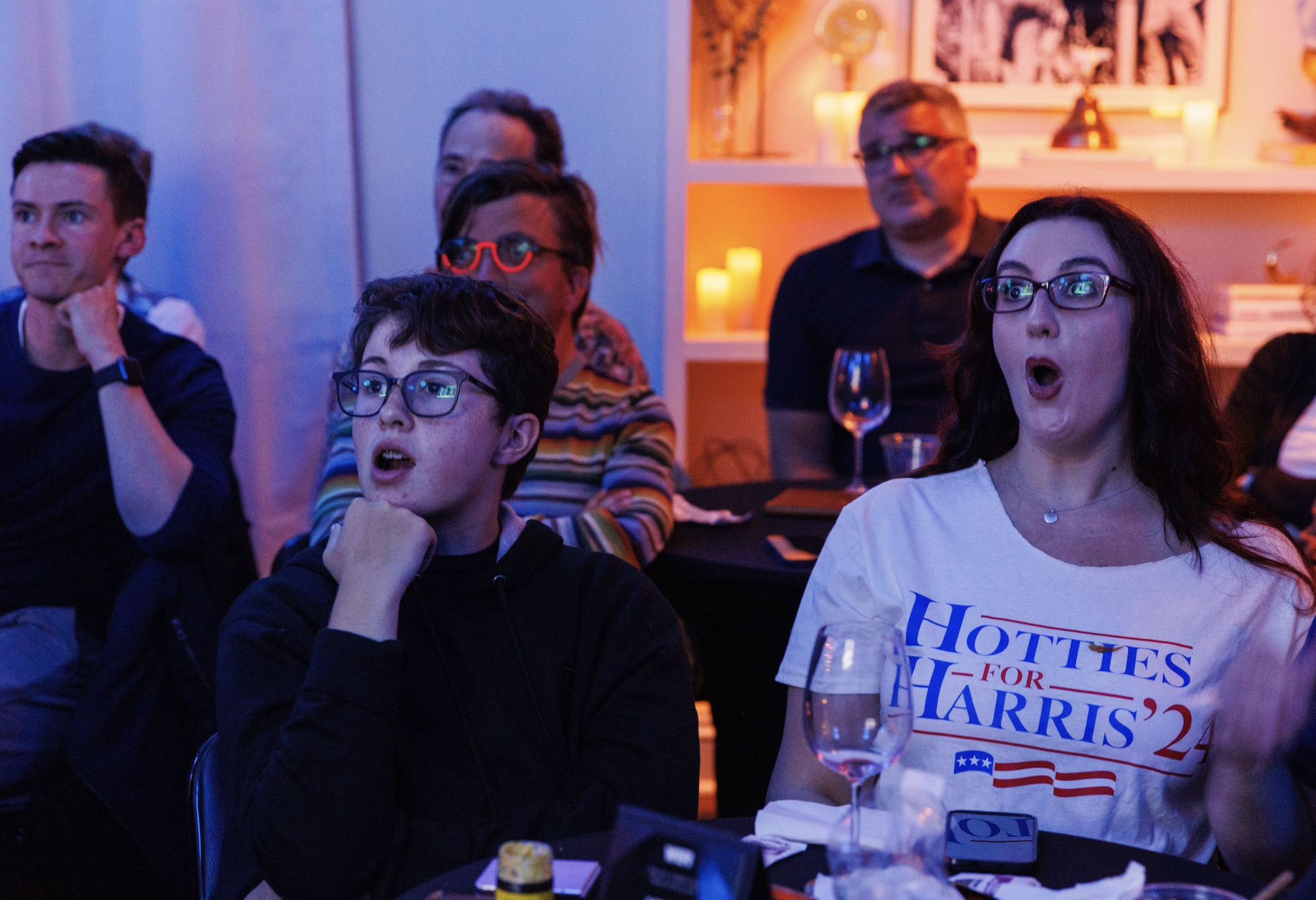 A group of people sit at a table with surprised expressions. One person wears a &quot;Hotties for Harris '24&quot; t-shirt