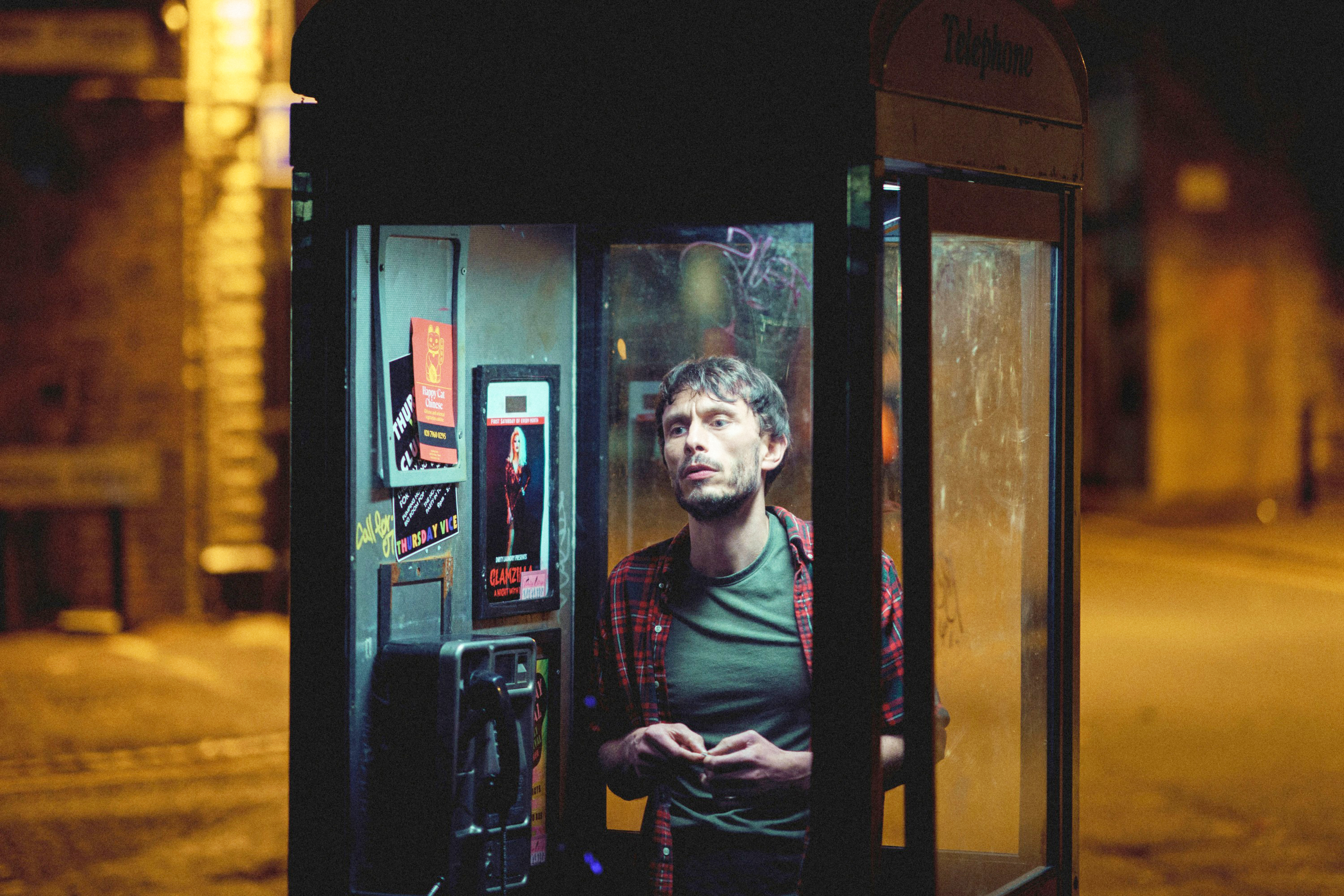 A man in a phone booth at night, wearing a T-shirt and a plaid shirt, stands under a streetlight, with s visible on the booth's interior walls