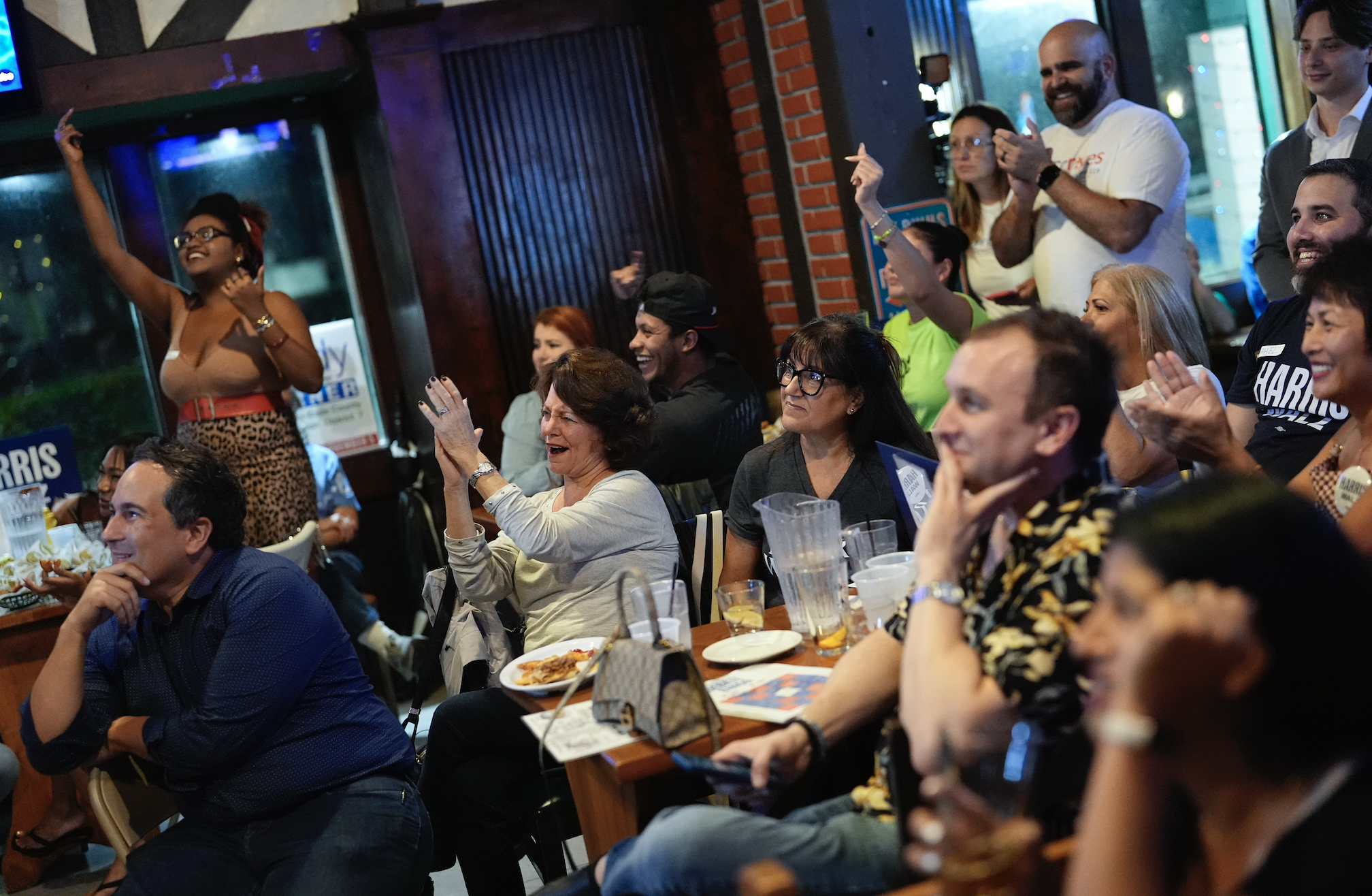 A group of people gather at a bar, clapping and cheering while watching an event. Some are seated at tables with drinks and food