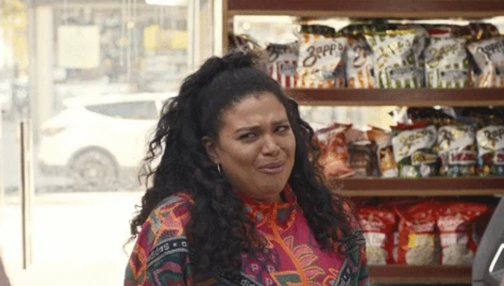 A woman with long curly hair styled in a high ponytail reacts with a bashful expression. She stands in front of a display of various snack bags