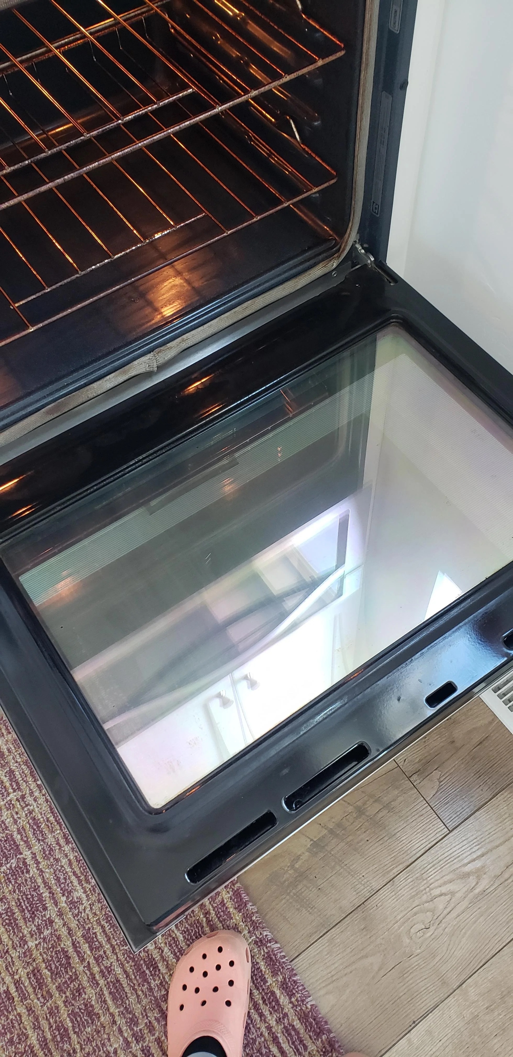 Open oven door with a reflective glass surface, showing the interior of an empty oven. Person wearing pink Crocs standing in front