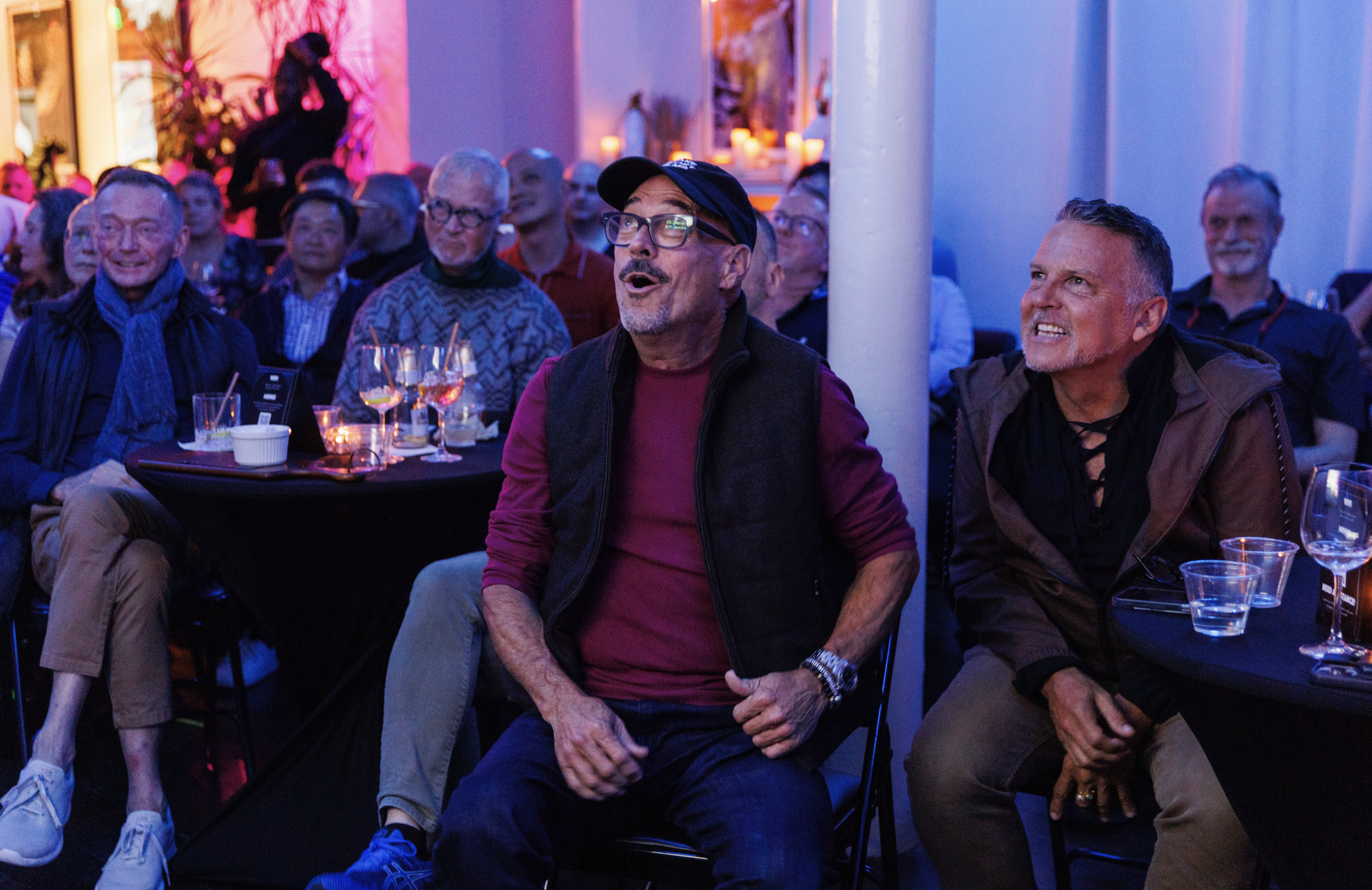 A group of people attending an event sit around tables, watching and reacting with smiles and expressions of surprise