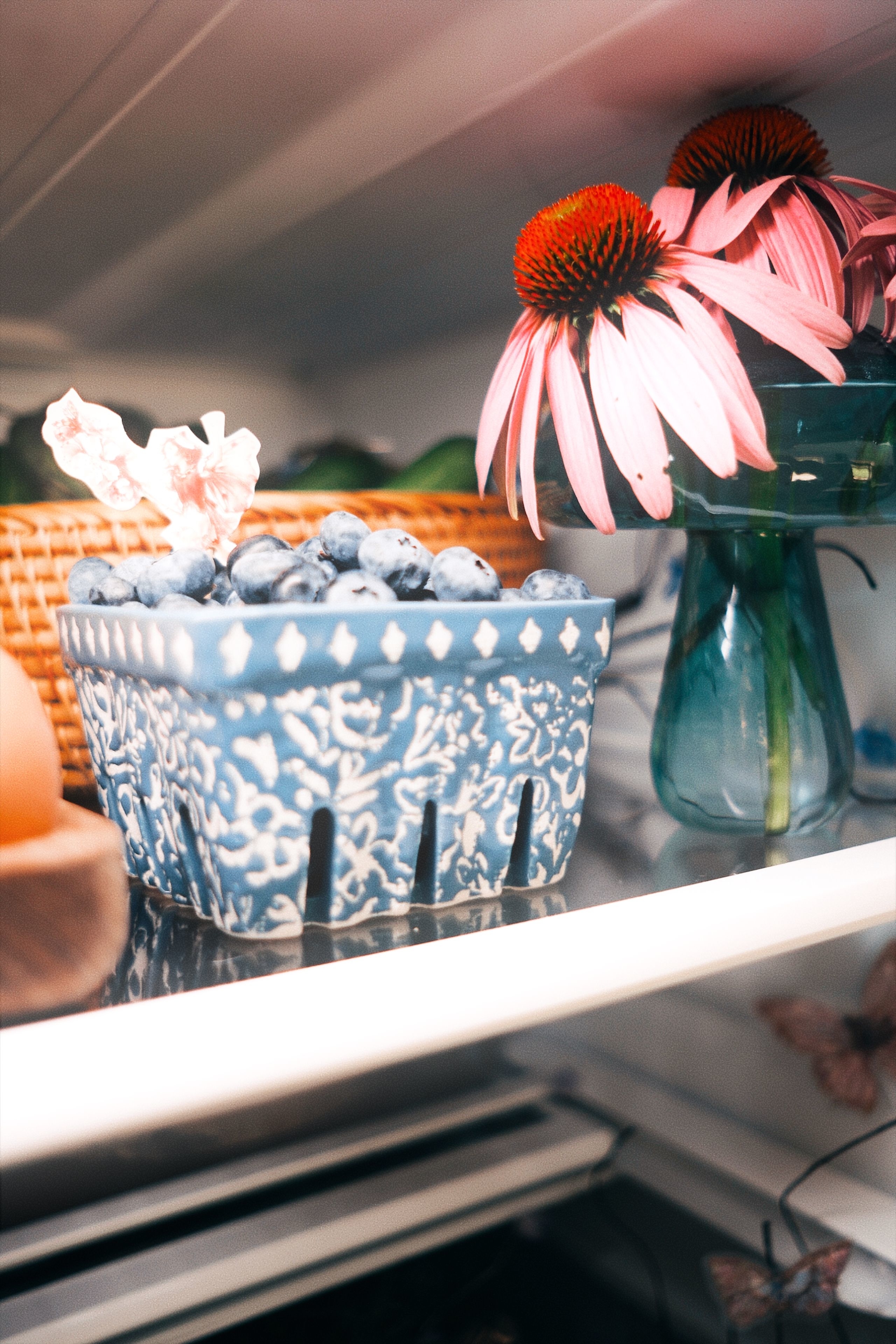 A ceramic container filled with blueberries is on a refrigerator shelf next to a glass vase with pink flowers