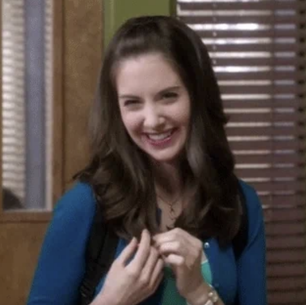 A person with long hair smiles while holding their backpack straps, standing indoors, possibly in a classroom or office setting