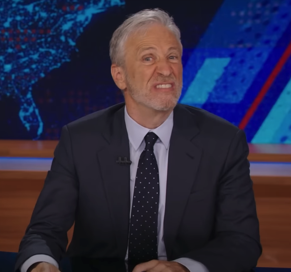 Jon Stewart at a news desk, wearing a suit and tie, making a humorous expression