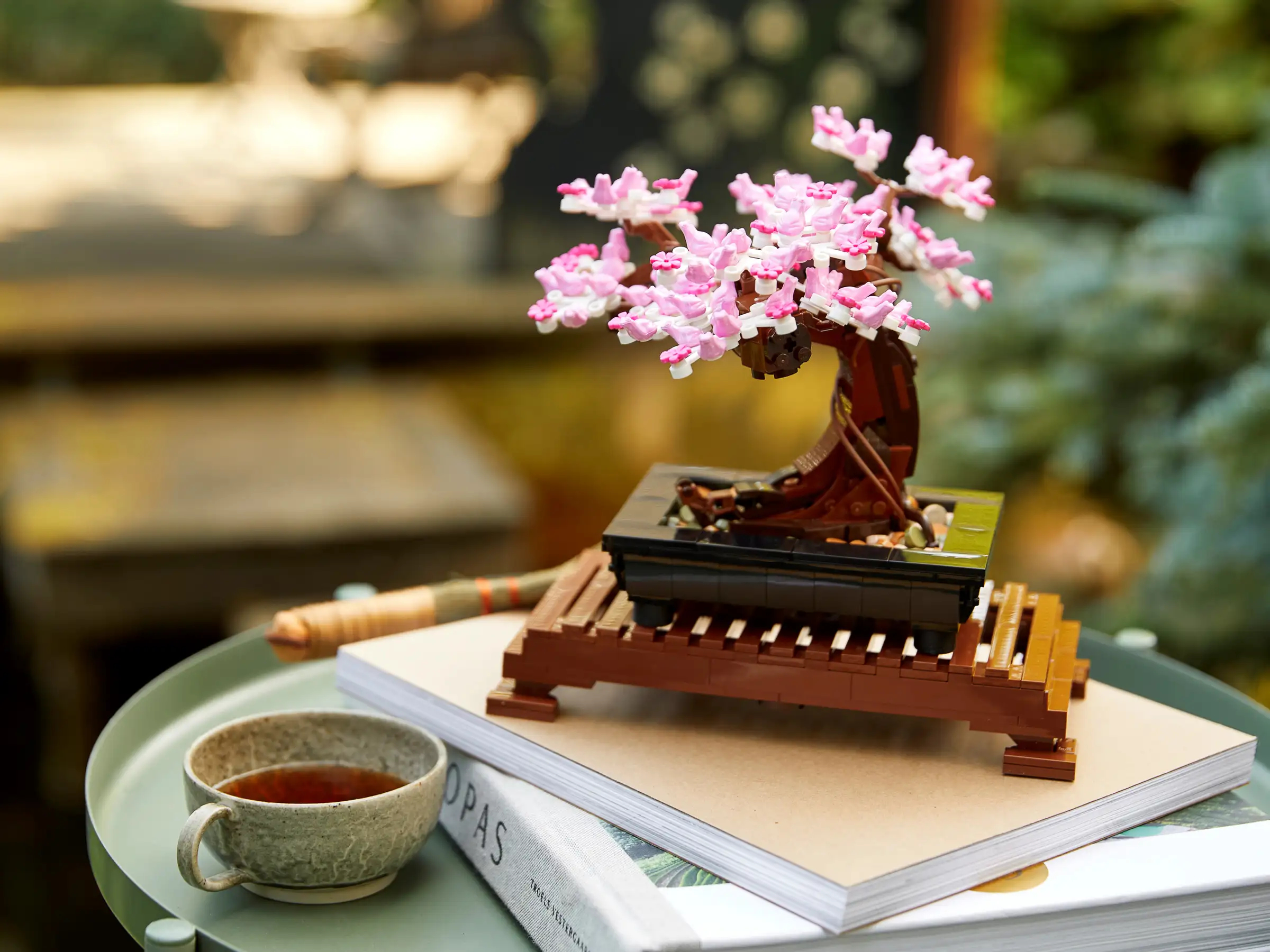 Pink floral model tree on a stand, placed on a table with a mug, a closed book, and a pen. Background shows a garden setting