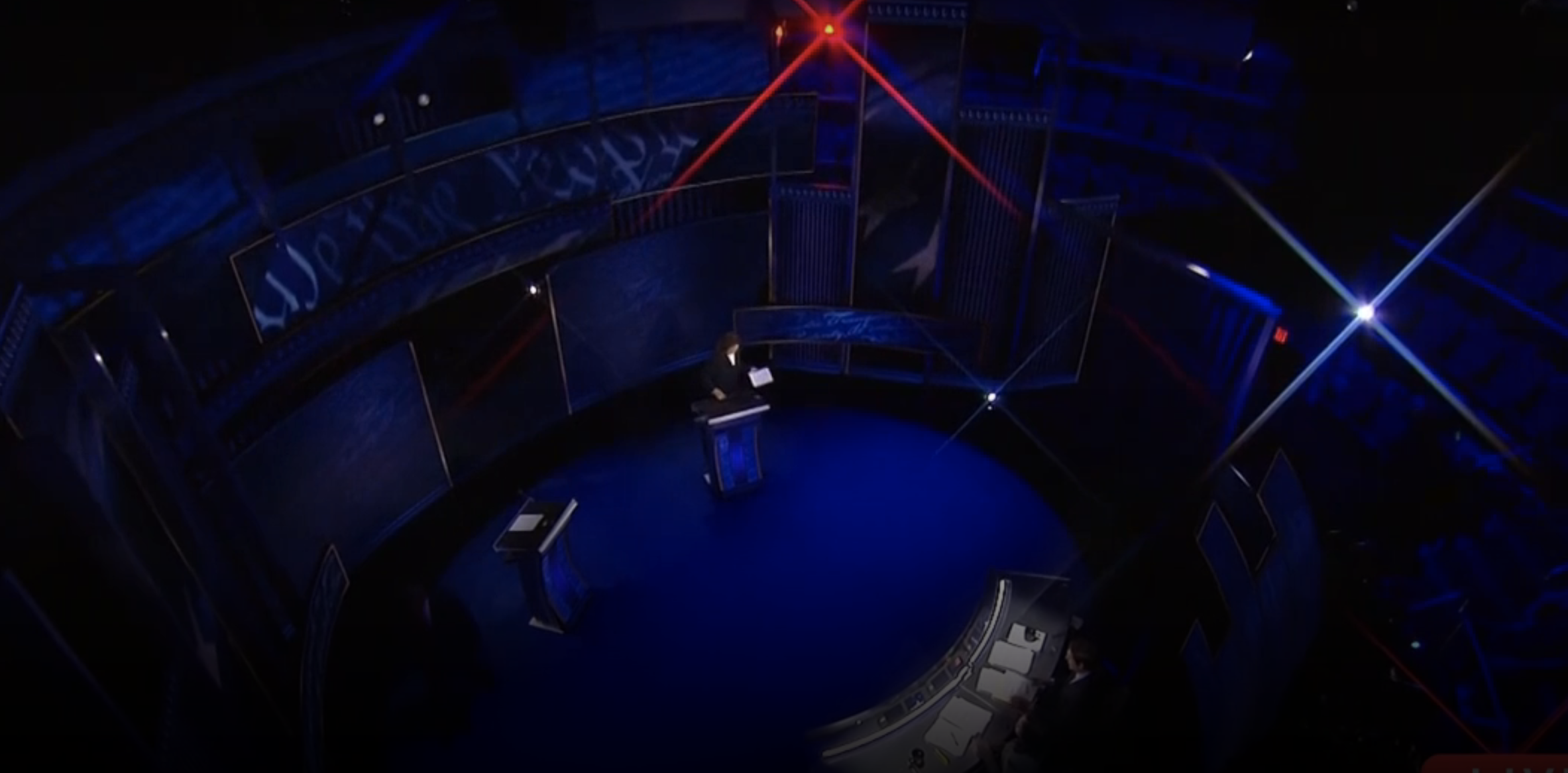 Wide-angle shot of a debate stage with one standing speaker at the podium, and a panel of three judges seated at a desk in a darkened auditorium