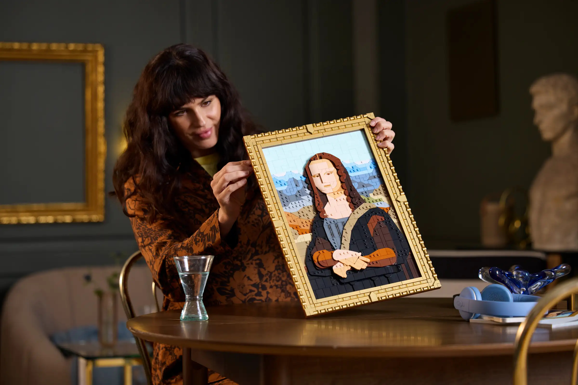 Woman holding a framed image of the Mona Lisa made from small building blocks, seated at a table with a glass of water and decor in the background
