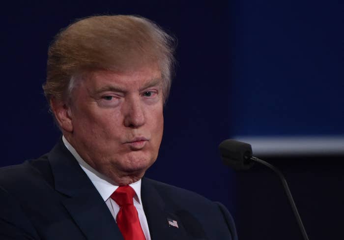 Donald Trump standing in front of a microphone, wearing a dark suit, white shirt, and red tie, with a concerned expression on his face