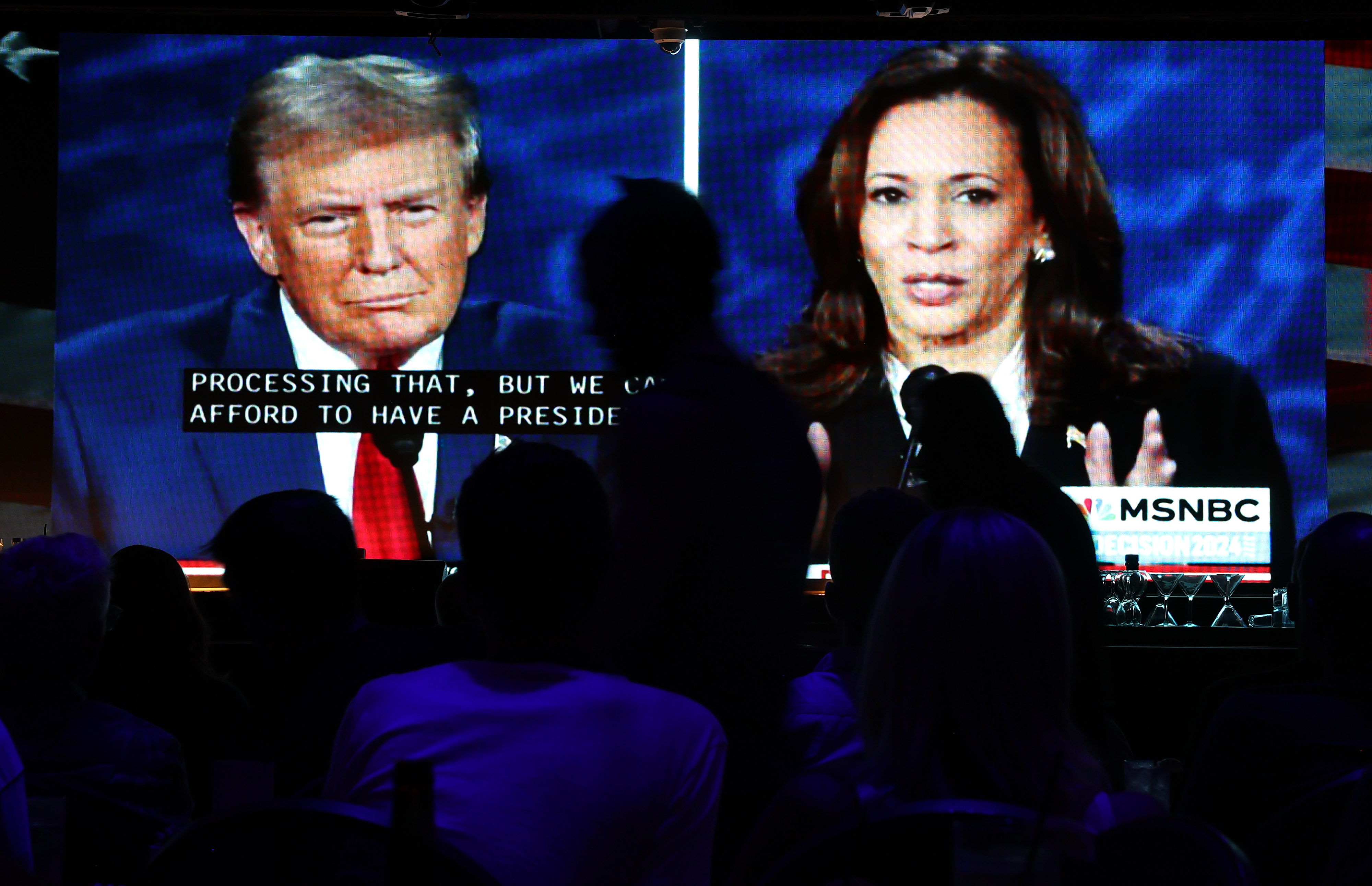 Donald Trump and Kamala Harris are shown speaking in separate frames on a TV screen during an MSNBC event. Silhouetted audience members watch the broadcast